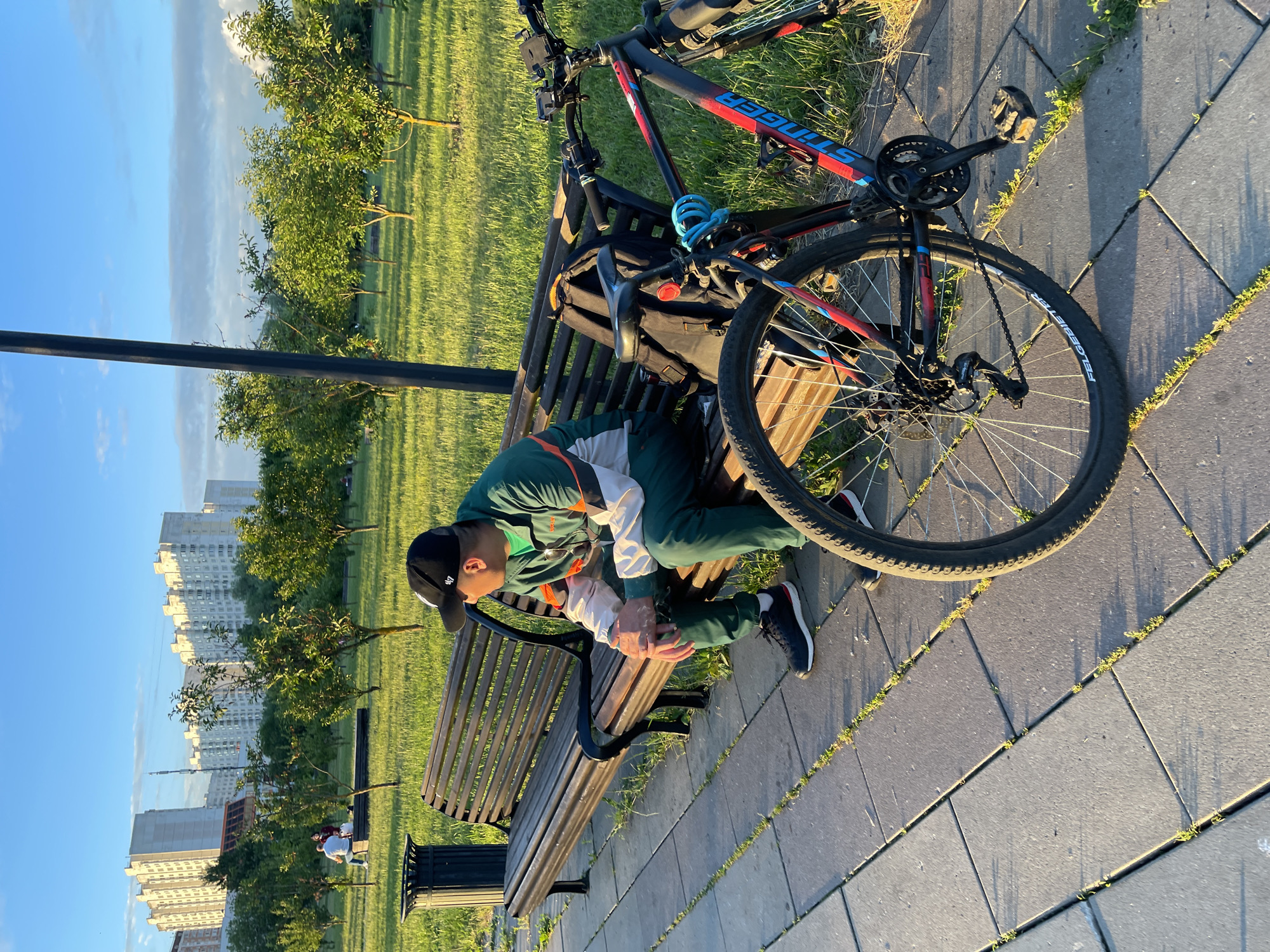 A person wearing a black baseball cap, green tracksuit, and black sneakers sits on a wooden park bench, looking to their right. A bicycle with a black frame, red and blue accents, and a blue U-lock is parked beside the bench, leaning against its back. A black backpack rests on the bench behind the person. The park features green grass, several small trees, and apartment buildings in the background. The scene appears to be in an urban park in Moscow, Russia, under a clear blue sky with some clouds. The ground is paved with grey tiles, and a dark trash can is visible to the left of the bench. The lighting suggests it is late afternoon or early evening.