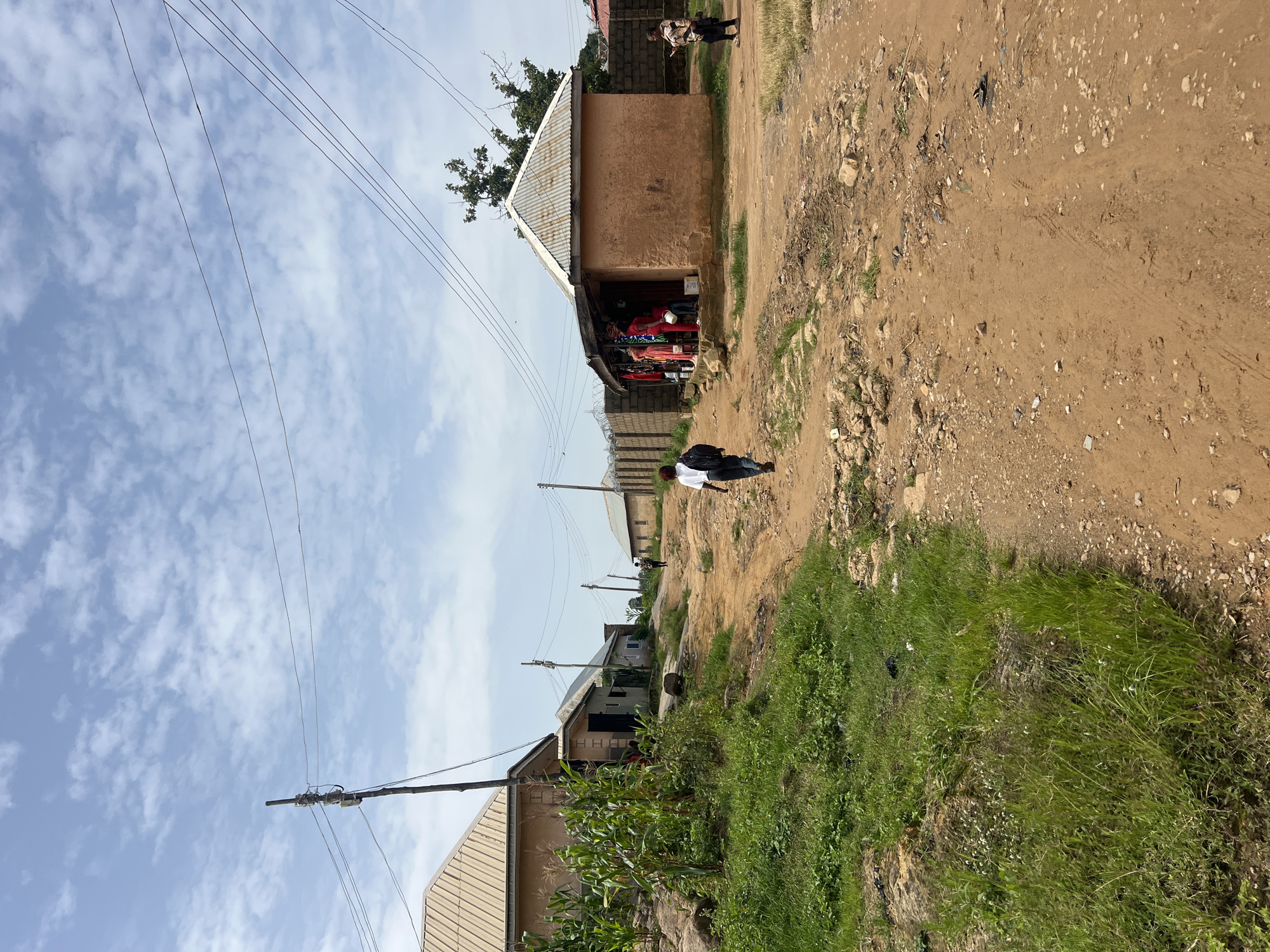 The image depicts a dirt road in a residential area of Jos, Nigeria. The road is unpaved and appears to be made of reddish-brown soil, with rocks and debris scattered across it. On the left side of the road, there are several buildings, including a house with a corrugated metal roof and a cornfield growing nearby. On the right side of the road, there is a small shop with a corrugated metal roof and an open front, displaying clothes for sale. Several people are visible in the image: a person walking away from the camera on the dirt road, another person standing in front of the shop, and two individuals further down the road, one of whom is walking away. The environment is somewhat overgrown with grass on the sides of the road, and there are utility poles with power lines running overhead. The sky is partly cloudy, indicating a daytime scene.