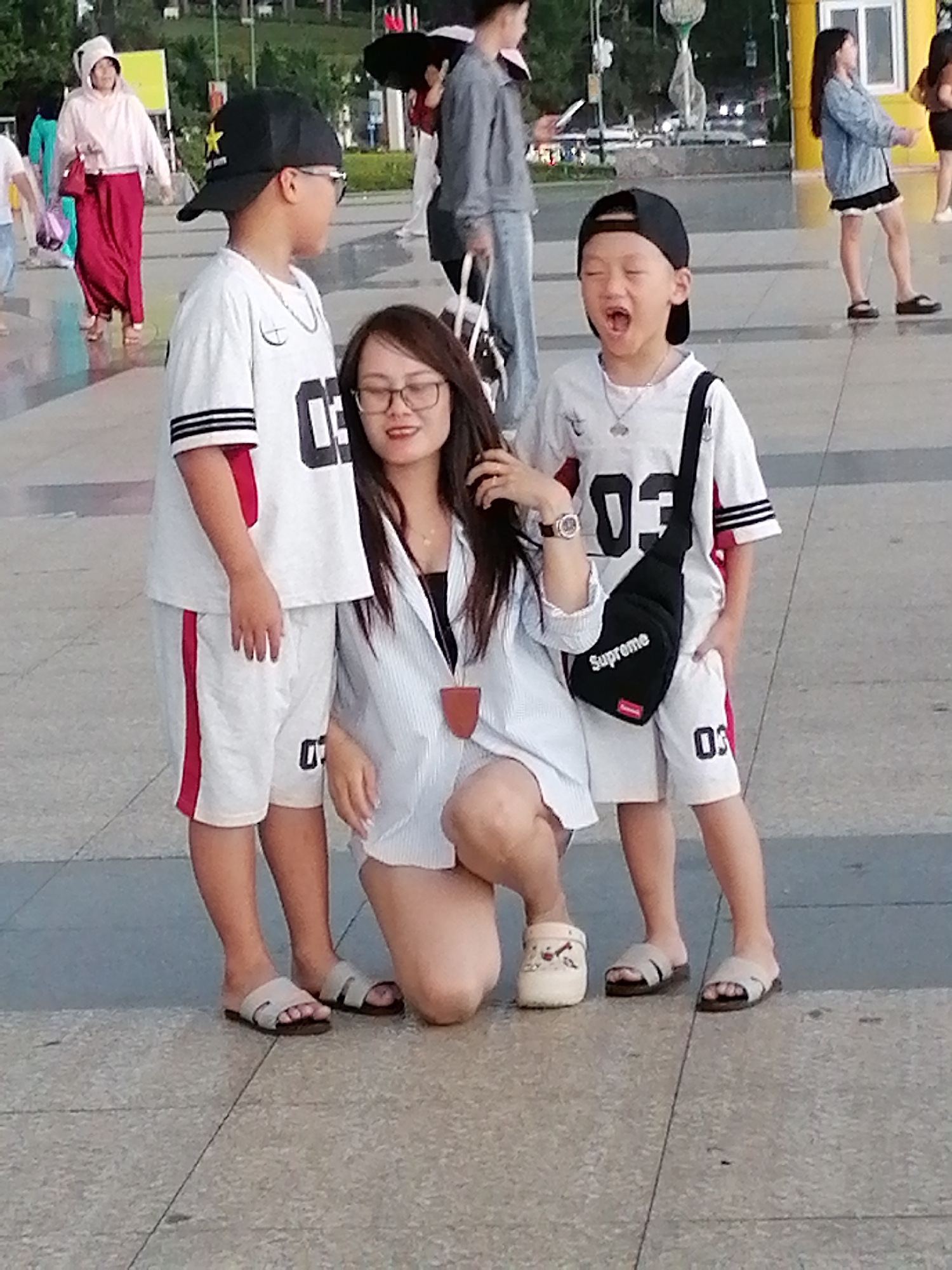 This is a medium shot of a woman and two young boys on a paved pedestrian area in Da Lat, Vietnam. The woman is kneeling and wearing a light blue pinstriped shirt and white shorts. She has glasses on and is looking at the camera. A young boy on the left is standing and wearing a white shirt with red trim and white shorts with red trim. He has a black baseball cap and glasses on and is looking to his right. A young boy on the right is standing and wearing a white shirt with black and red trim and white shorts with red trim. He has a black baseball cap on and is looking forward with his mouth open. He is also wearing a black crossbody bag with "Supreme" written on it. In the background, other people are walking in a park-like setting.