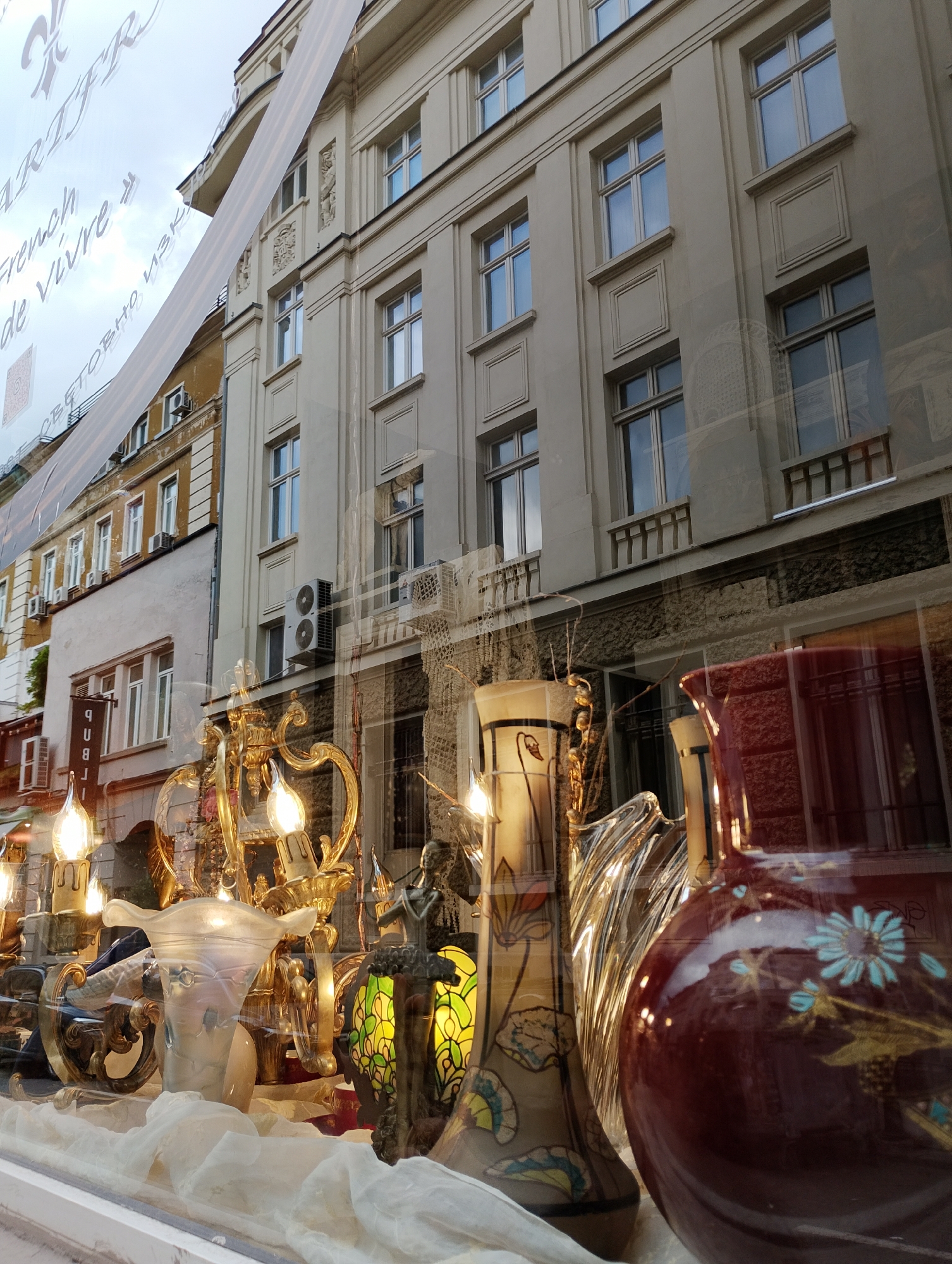 The image displays a shop window, presenting a complex visual layering of items inside the store and reflections of the urban environment outside.
Inside the shop window, a curated display of decorative objects is arranged on white, draped fabric. In the foreground, a prominent, dark red ceramic jug or vase is visible, decorated with light blue floral patterns and subtle gold accents. To its left, a tall, slender vase features art nouveau-style botanical motifs in muted green, yellow, and brown. Numerous ornate, golden candelabras or light fixtures with exposed, flame-shaped bulbs cast a warm, inviting glow across the display. Other items include a clear, fluted glass object, a frosted white glass vase with a flared rim, and a small, classical figurine positioned next to a lamp with green stained-glass-like patterns.
The reflections on the shop window reveal a multi-story, light-colored building with an older architectural style, characterized by numerous rectangular windows, some with decorative surrounds and small balconies. Beyond this, further reflections show older, less ornate buildings, one yellow and one white, both with several air conditioning units attached to their facades. An overcast sky is reflected at the top of the window.
Visible text on the window, appearing reversed due to reflection, includes partial English phrases like "French" and "de Vivre" (likely part of "Art de Vivre" or "Joie de Vivre"). Cyrillic script is also visible, reading "СВЕТОВНО ИЗК," which likely refers to "СВЕТОВНО ИЗКУСТВО" (World Art). A partial reflected sign on a building in the background reads "PUB." The scene takes place during an overcast day, suggesting an urban setting, possibly in an older European city given the architecture and Cyrillic script.