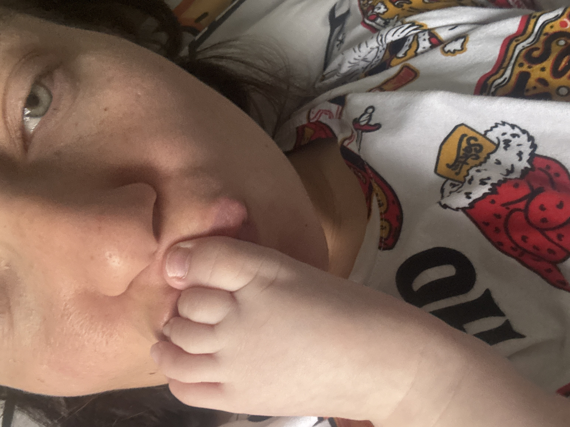 The image captures a close-up, intimate moment between an adult and an infant, likely indoors in Раздолье, Russia. The left side of the frame shows a partial view of an adult's face, featuring a light-colored eye (green/hazel), nose, and cheek, with dark hair visible. The right side and foreground are dominated by a baby's small, pale foot, with visible toes and fingernails, gently pressed against the adult's face, specifically near their upper lip and nose area.
The adult is wearing or is against a white fabric patterned with various designs in red, black, and gold/yellow. The pattern includes stylized figures, one resembling a royal character with a fur-trimmed robe and crown, holding a gold plaque with some indistinct text (possibly "SOT"). Other graphic elements and large, partially visible black text that appears to include the letter "O" or "IO" are also present.
The soft, ambient lighting suggests an indoor setting. The scene conveys a strong sense of warmth, affection, and a loving bond between the adult and the baby, highlighting a tender interaction. No specific time of day or other location cues are discernible from the image itself beyond the provided context.