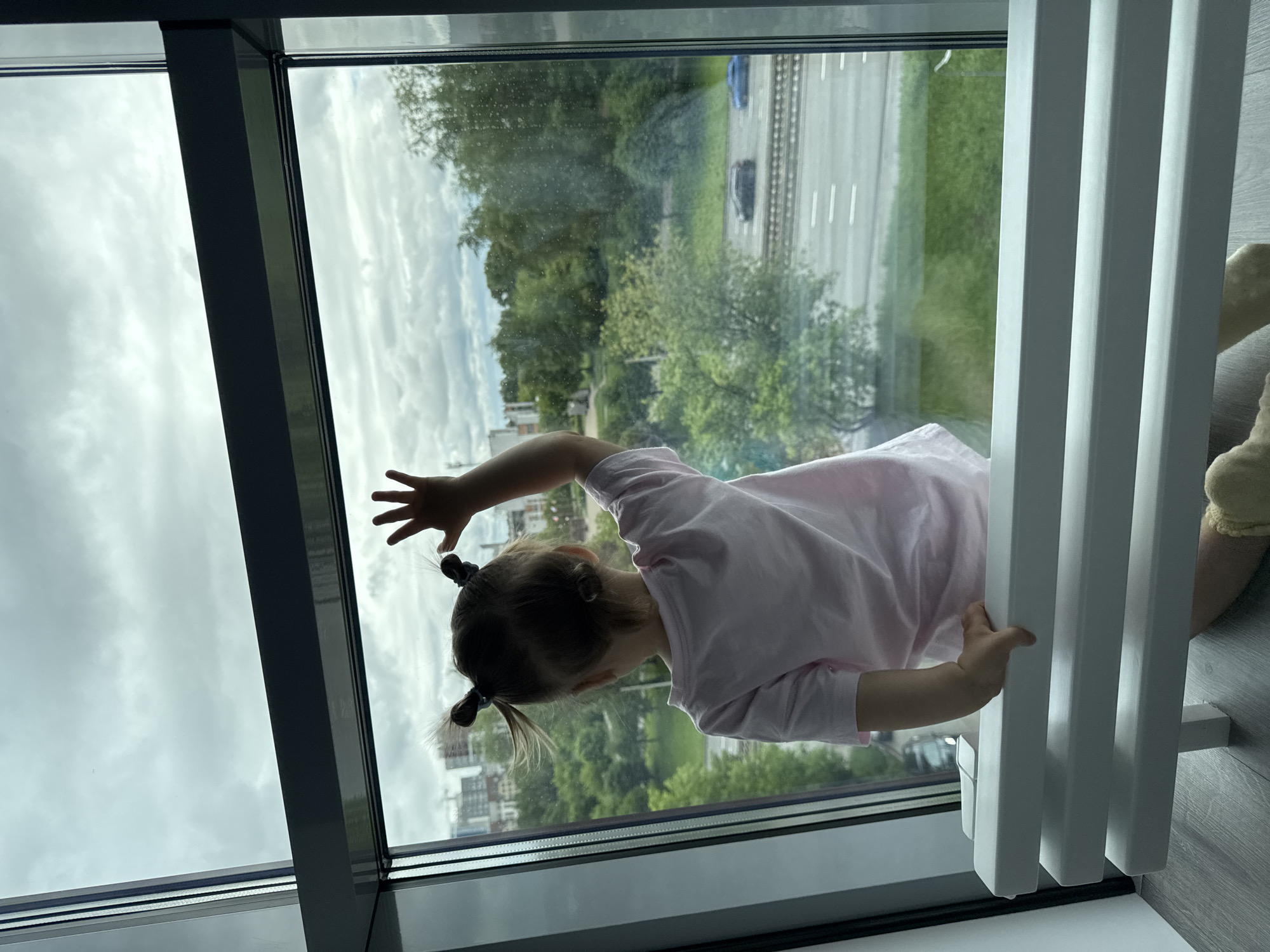 A young child, facing away from the camera, is pressed against a large window overlooking a cityscape. The child, wearing a light pink shirt and with their hair styled in two pigtails secured with blue fasteners, has one hand pressed flat against the glass. Outside the window is a view of Izhevsk, Russia, including buildings, green trees, and a multilane highway with moving vehicles. A white radiator stands near the child on the right side of the image. The sky is overcast with grey clouds.