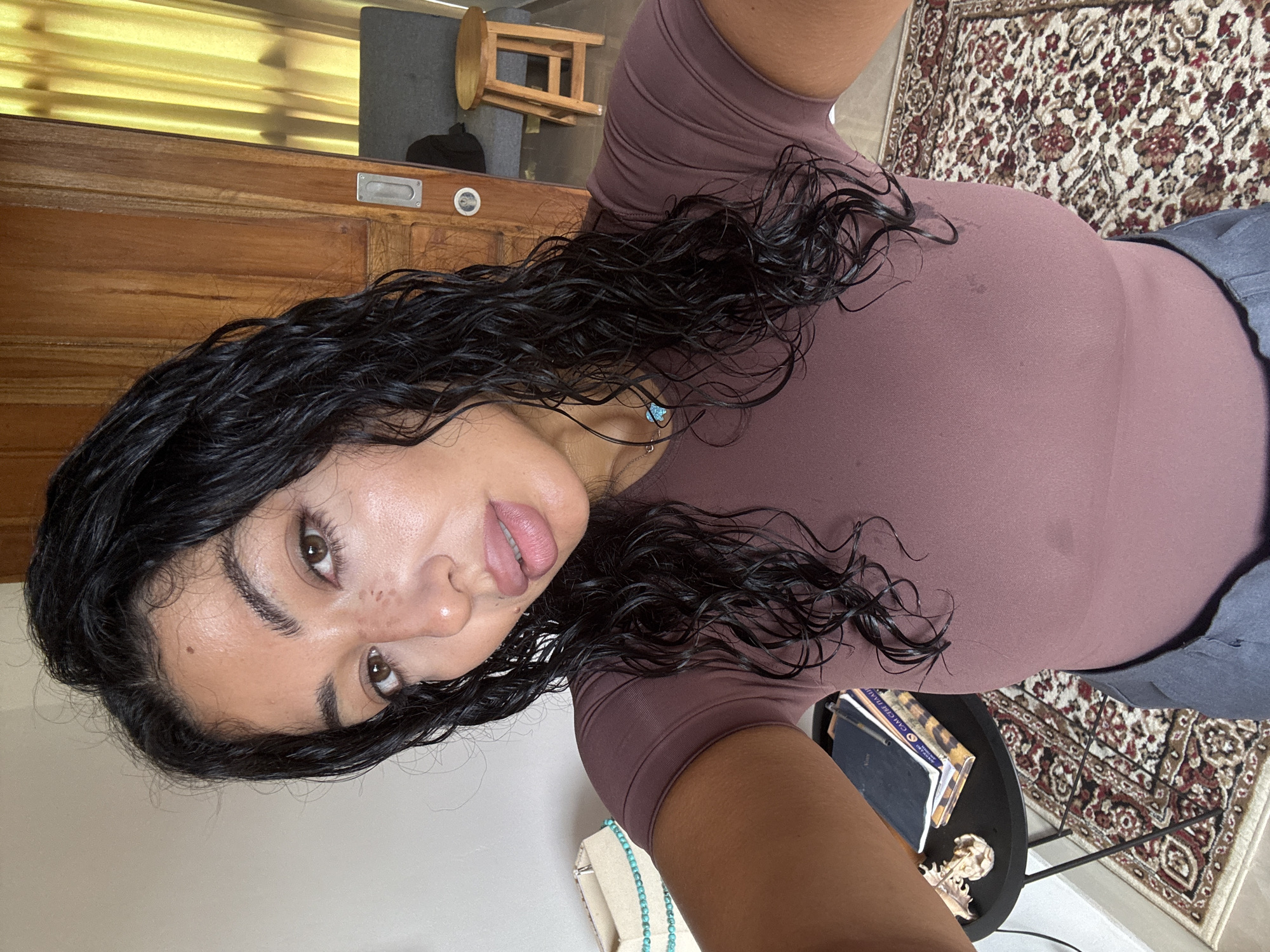 A selfie from an overhead angle of a woman indoors in a room in North Kuta, Indonesia, based on cultural indicators. The woman has long, dark, wet, curly hair and olive skin, wearing a mauve-colored top. A turquoise necklace is visible around her neck. Behind her is a wooden door, a rug, and a small round table. Books and a decorative skull are on the table.