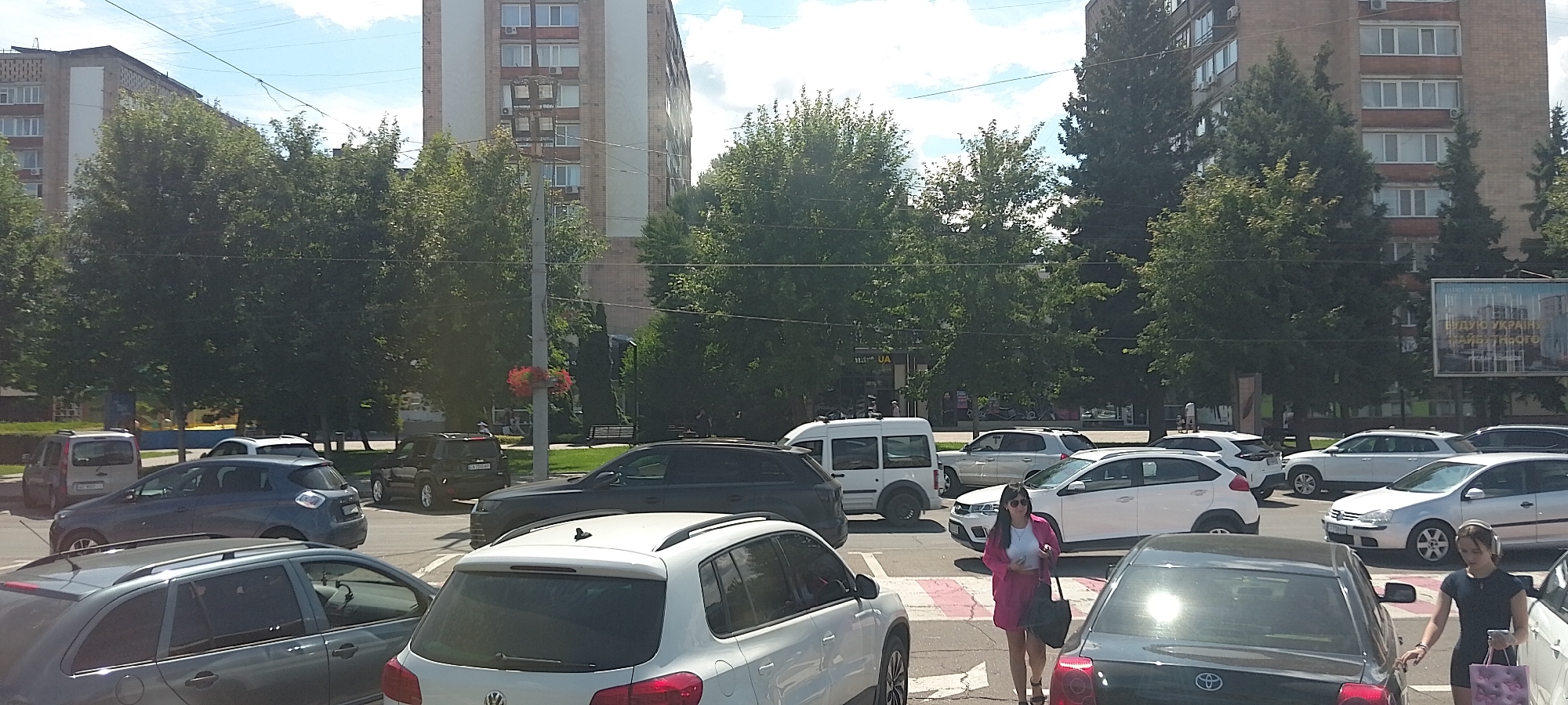 The image depicts a street scene in Cherkasy, Ukraine, with several cars parked and in motion. In the foreground, a dark grey car is partially visible on the left, and a white Volkswagen hatchback is in the center. To the right of the Volkswagen, a black car is positioned. A pedestrian, a young woman wearing headphones, is walking near the black car. Behind these vehicles, a variety of cars are visible, including a white compact SUV, a dark grey SUV, and a silver sedan. Further back, a white van is parked. Several trees line the street, and apartment buildings are visible in the background, one of which has a billboard advertising a future Ukraine. The sky is partly cloudy.