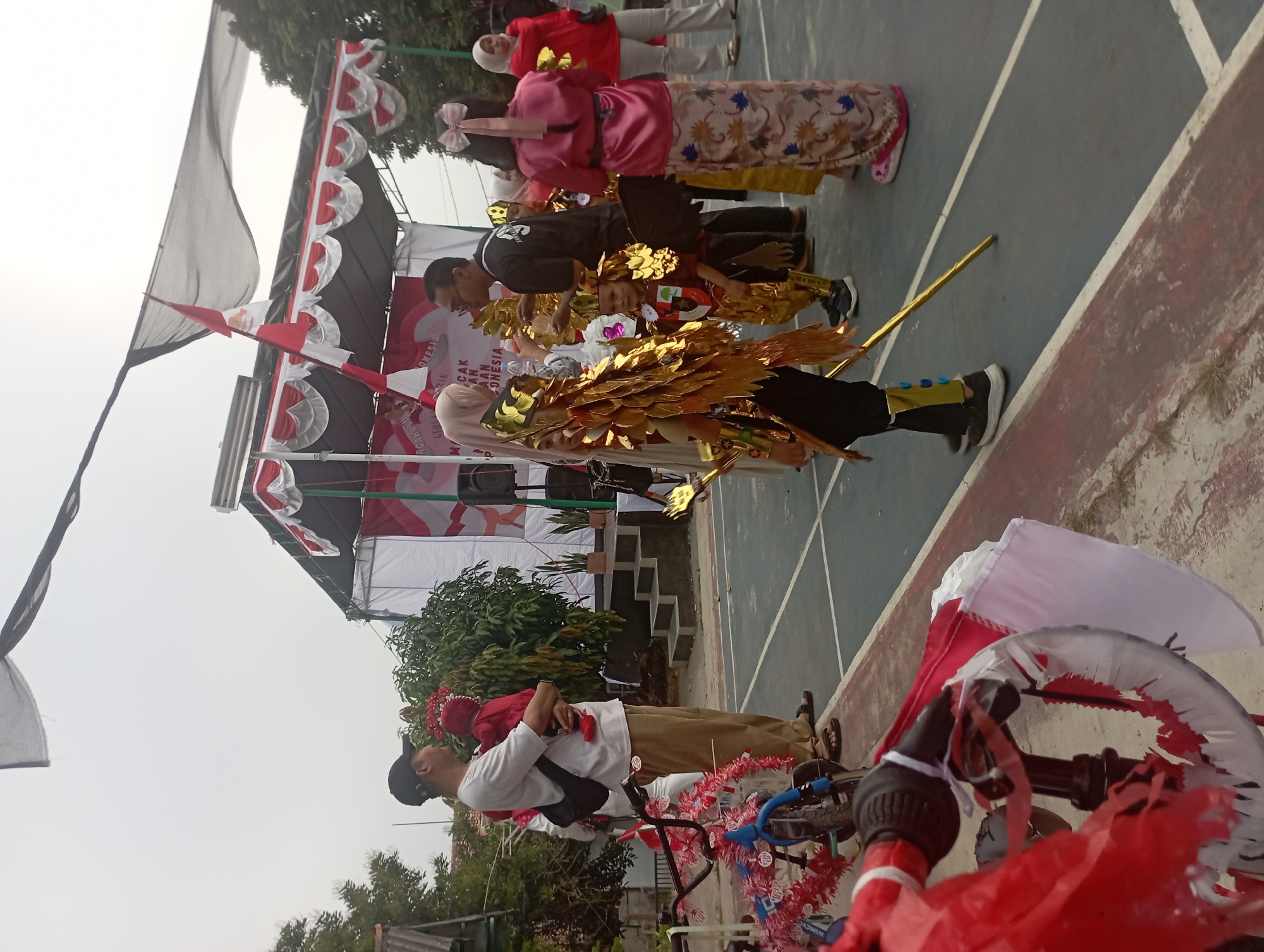 In Purwakarta, Indonesia, a daytime outdoor scene depicts a gathering with a stage in the background. A man wearing a white t-shirt, brown pants, and a black cap holds a baby dressed in a red outfit. Several children in elaborate, gold-colored costumes, some with wings, are positioned in the foreground. One child holds a golden staff. A woman in a pink outfit with a large bow in her hair stands to the right, facing away from the camera. Other individuals, including a woman in a red top and another in a headscarf, are visible. A bicycle decorated with red and white streamers is in the lower left corner. The ground appears to be a sports court. The sky is overcast.