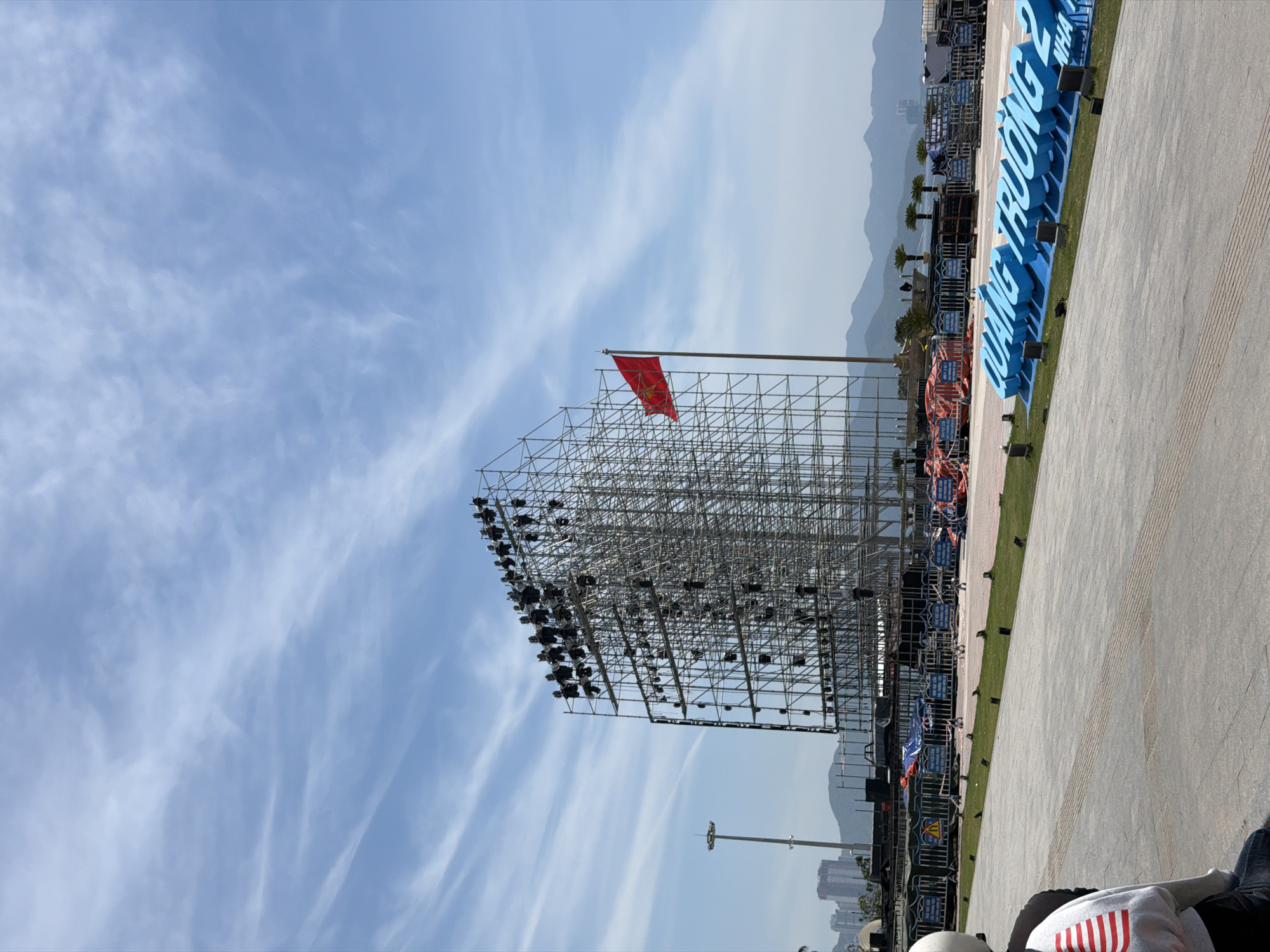 The image captures a wide outdoor scene with a bright blue sky and scattered white clouds. In the foreground, a paved area leads to a grassy strip where large, three-dimensional blue letters spelling "QUANG TRUONG" are displayed, along with partial text indicating "NHA". Beyond the grass, a large metal scaffolding structure is erected, covered with numerous lights. A red flag with a yellow star, likely the Vietnamese flag, flies from a pole to the right of the scaffolding. In the background, hills and mountains are visible, with some buildings on the left. Several barriers and orange tarpaulins are present around the scaffolding area, suggesting event preparation. A person wearing a white helmet and a grey and white striped shirt is partially visible on the left edge of the frame, sitting on what appears to be a motorcycle. The overall scene suggests an outdoor event or festival setup in Nha Trang, Vietnam.