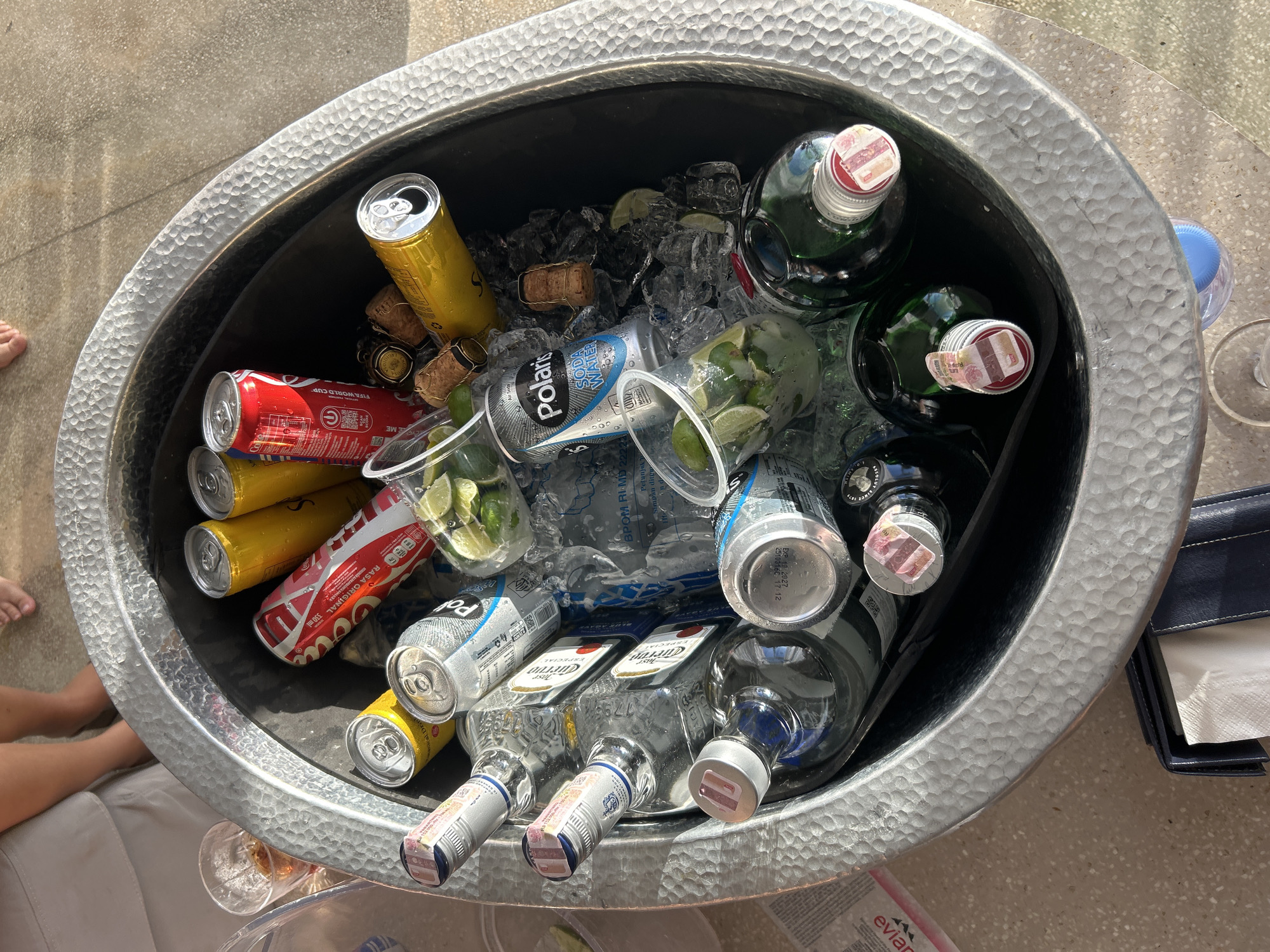 A top-down, medium shot captures a hammered metal tub filled with ice and beverages, likely at an outdoor setting in North Kuta, Indonesia. The tub contains several bottles of clear liquor, distinguished by silver and blue labels, and some green bottles with red-capped tops. Also visible are cans of Coca-Cola, yellow cans, and cans labeled "Polaris Soda Water." Several plastic cups, each containing lime wedges and ice, are scattered among the drinks. Two corked wine bottles, likely champagne, are partially submerged in the ice. The blurred background shows a person's legs and feet, suggesting they are sitting or standing near the tub. The ground appears to be a textured, light-colored surface, possibly a patio or walkway.