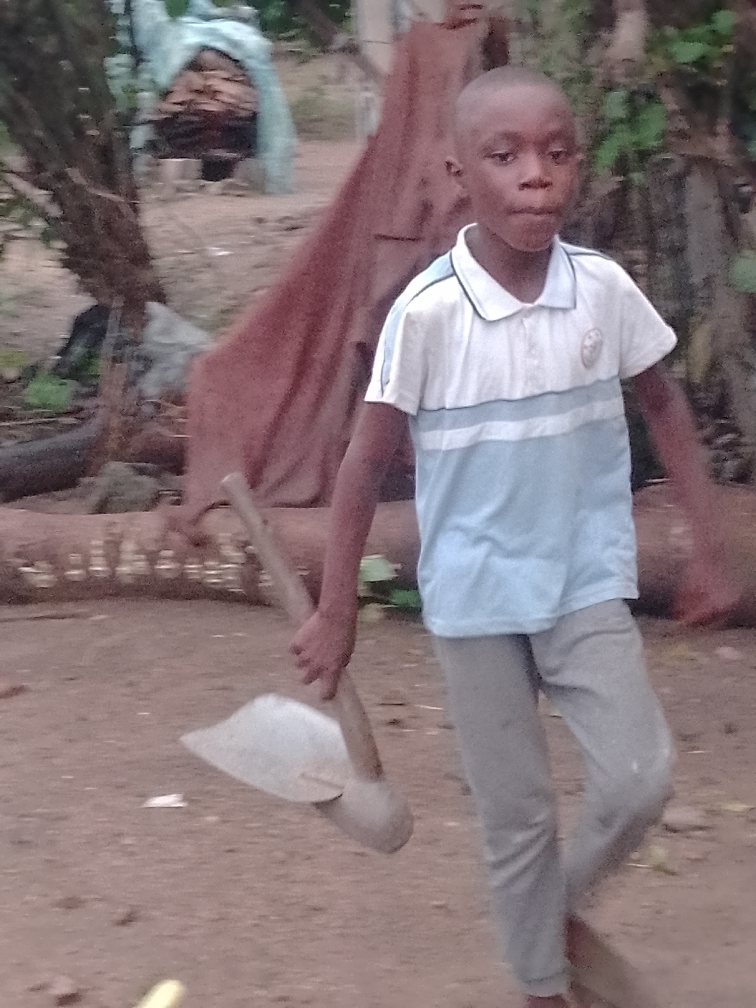 A young boy is walking with a spade in the location of Unknown, Unknown. He is wearing a blue and white shirt and gray pants. The environment appears to be a dirt or gravel area with foliage and a tarp-covered structure in the background.