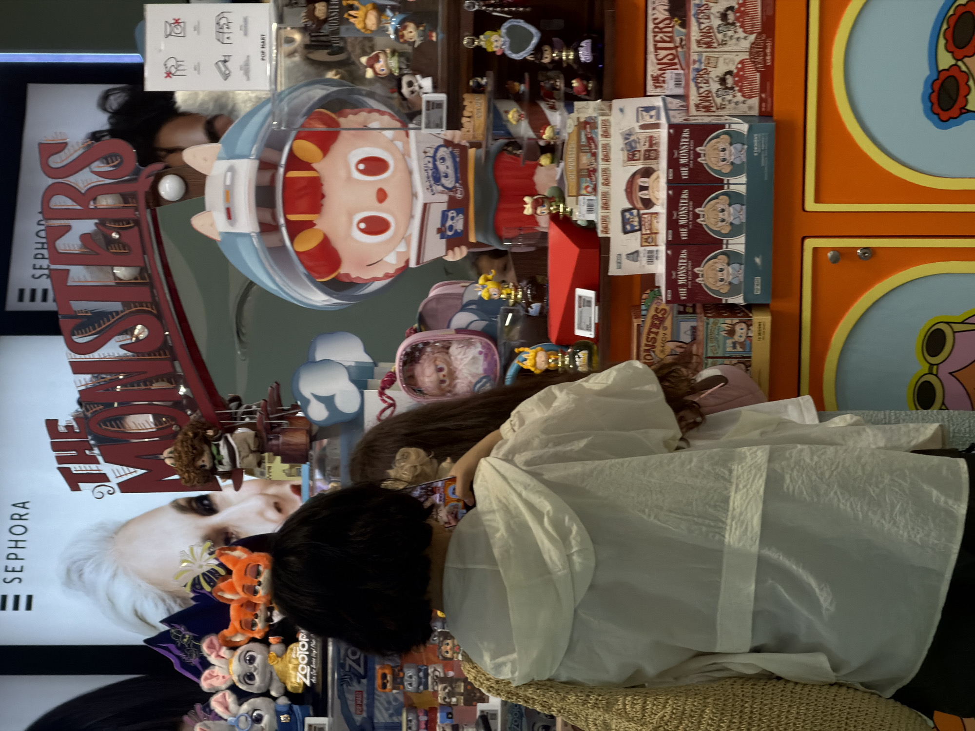 In a retail environment, likely a toy store in Wichit, Thailand, two individuals are partially visible, interacting with displayed merchandise. The foreground is dominated by the back of a person wearing a white hooded jacket and a woven straw bag. To their right, another person with long, dark hair is also visible, holding a smartphone. They are both looking towards a display of collectible toys. The display features various figures, including plush toys, miniature figurines in boxes, and larger character figures. Prominently displayed is a large, stylized character with a blue helmet and red hair, holding a book. Signage in the background includes "SEPHORA" and "THE MONSTERS," suggesting a themed promotion. The overall scene is well-lit, with a focus on the colorful array of toys.