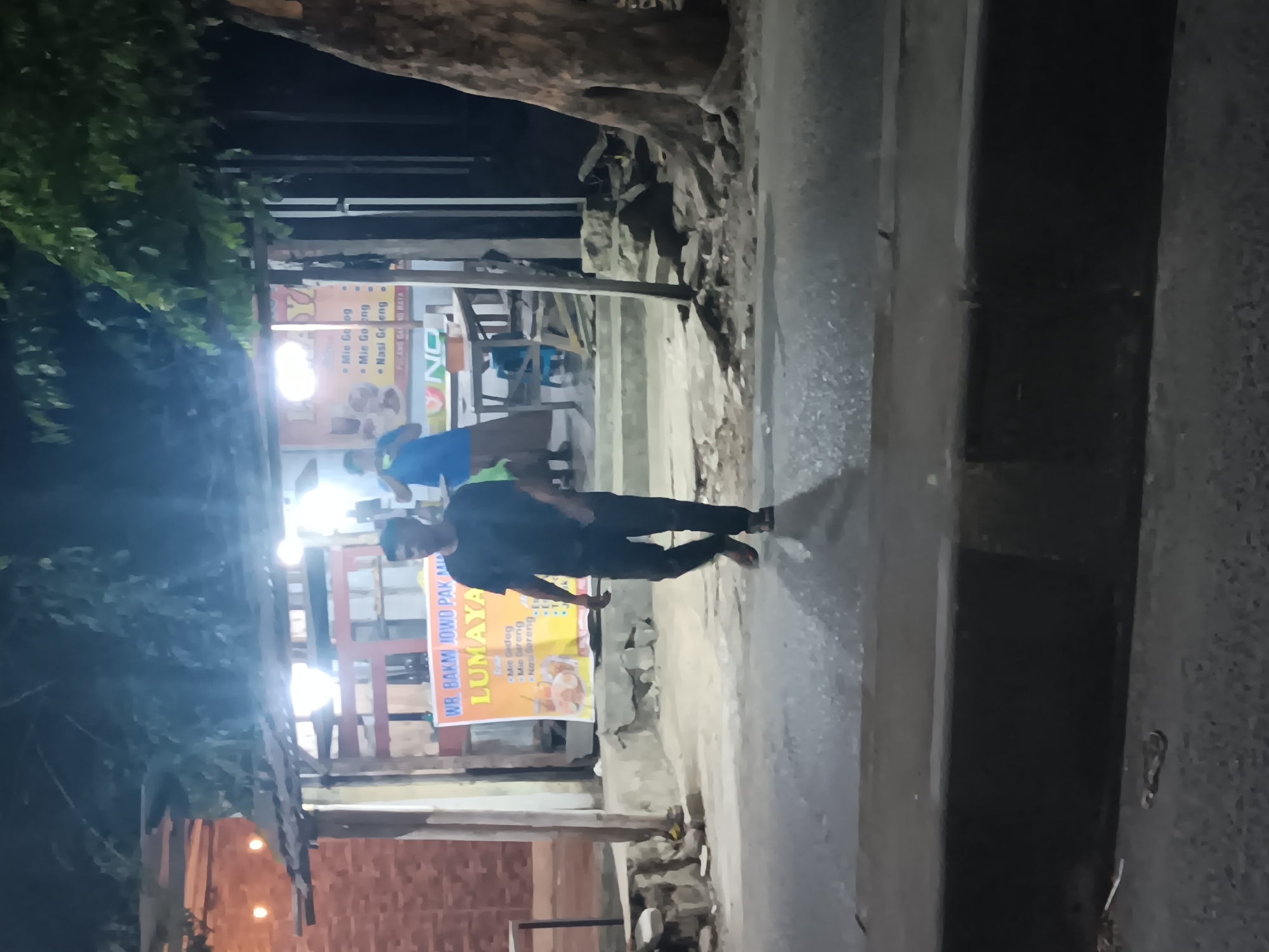 The image depicts an outdoor scene at night in Batursari, Indonesia, likely near a food stall. A man wearing a dark t-shirt and dark pants is walking from right to left across a paved road. Behind him is a small, open-air food stall with a thatched roof and a bright orange banner that reads "WR BAKMI JOWO PAK MIN LUMAYAN". The banner also lists menu items such as "Mie Gedeg", "Mie Goreng", and "Nasi Goreng". Another person, wearing a blue shirt and a brown sarong, is visible inside the stall, standing near a table. The stall is illuminated by artificial lights. Trees with large roots are visible to the right of the stall, and a brick wall can be seen on the left. The ground in front of the stall appears to be concrete, and there is debris scattered around the base of the trees and the stall. The road in the foreground is wet and reflects the lights.