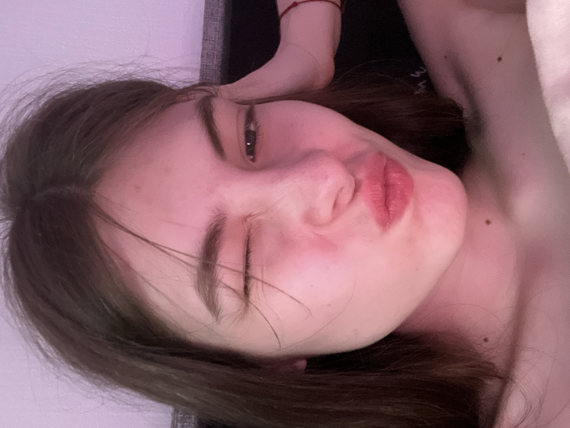 A close-up, tilted selfie shows a young woman with brown hair and fair skin. She is partially winking with her left eye and puckering her lips. Her expression is playful. A small red bracelet is visible on her left wrist, which is raised above her head. The background includes a dark, textured surface, possibly a headboard or wall, and a soft, light-colored material. The environment is ambiguous and does not provide clues for locating the city of Saint Petersburg, Russia.