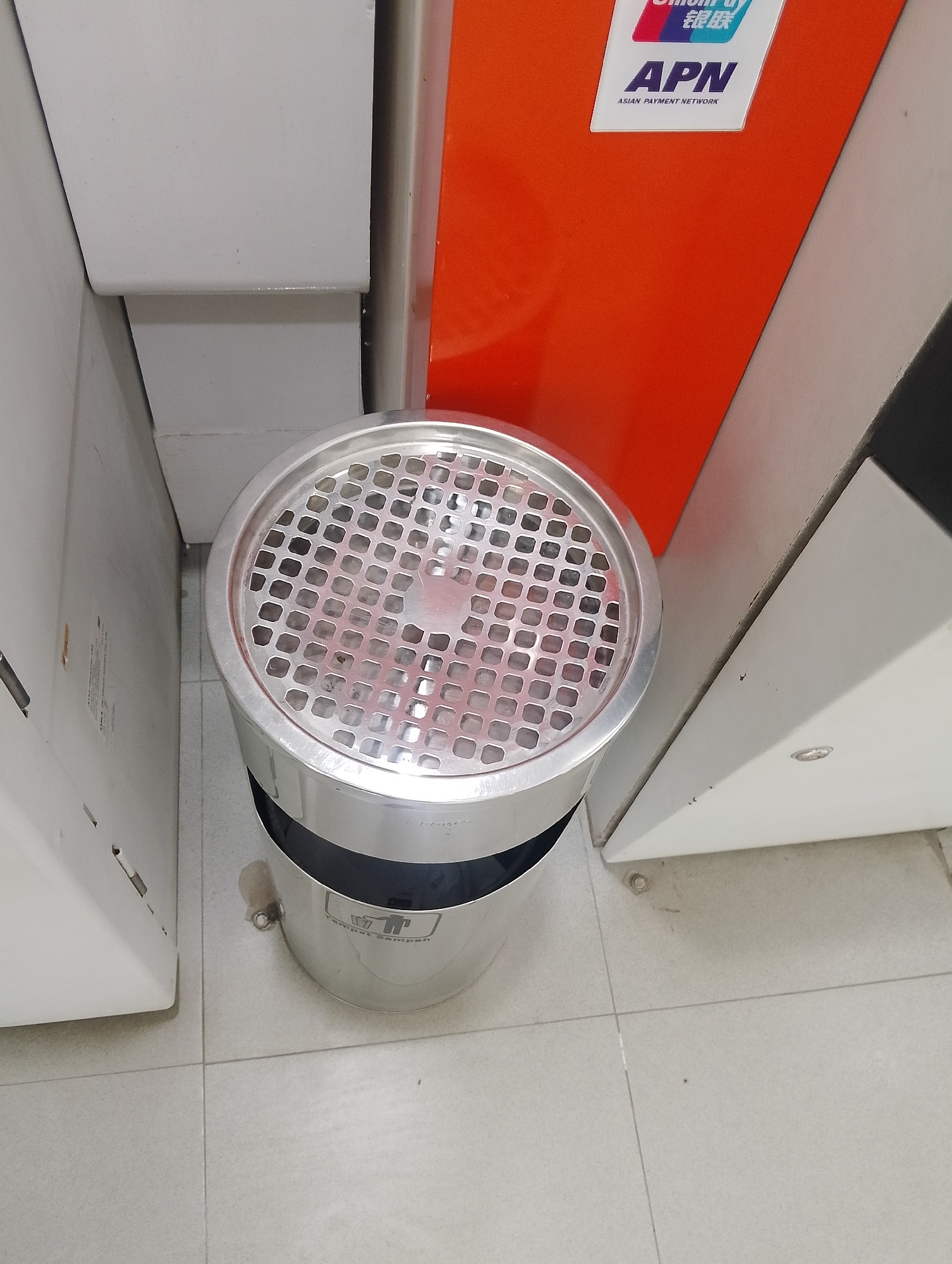 The image captures a street-level view of an outdoor area, likely a public space such as a roadside or walkway, in the city of Serbelawan, Indonesia. The primary subject is a stainless steel ashtray bin with a perforated lid, positioned centrally in the frame. The bin appears to be used, with remnants of cigarette butts visible through the perforations.
To the left of the ashtray bin, a portion of a white electronic device or kiosk is visible. Its surface is smooth and light-colored, with some visible wear and tear, including what appears to be a sticker or label. The upper part of this device features a gray, angled surface.
To the right of the ashtray bin, a large, bright orange panel dominates the background. A white sticker with a logo and text is affixed to this orange panel. The text on the sticker includes "APN" in large letters, with smaller text below it reading "ASIAN PAYMENT NETWORK." Above "APN," a smaller logo with Chinese characters and the text "UNION PAY" is partially visible. This orange panel appears to be part of a larger structure, possibly another kiosk or machine.
The ashtray bin is situated on a light-colored tiled floor, with visible grout lines. A dark shadow is cast behind the bin, suggesting an overhead light source or the presence of other structures not fully captured in the frame. The overall scene is utilitarian and suggests a functional outdoor space where smoking is permitted. The presence of the payment network logo might indicate proximity to a commercial establishment or a public transportation hub.