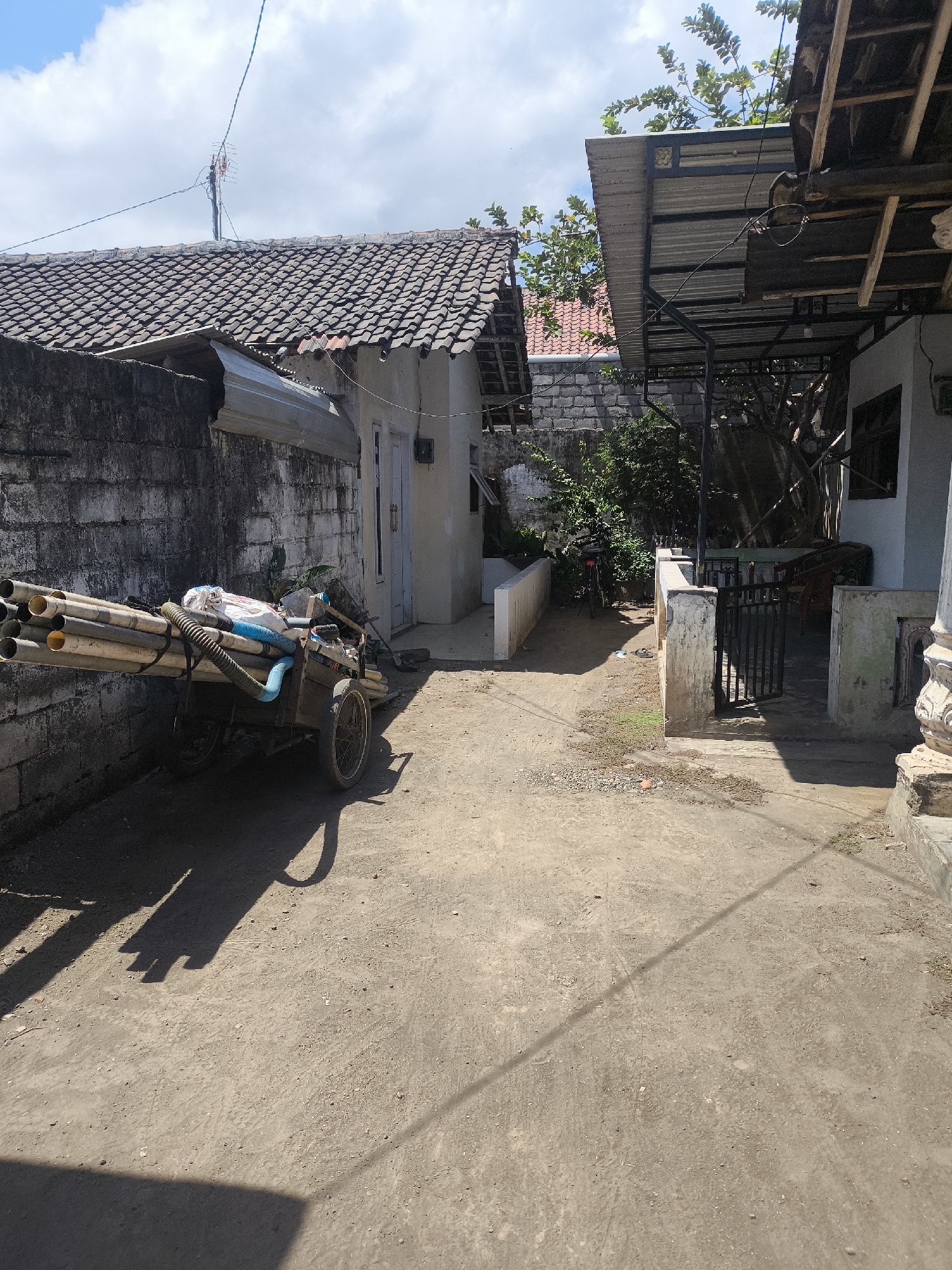 In Kebonsari, Indonesia, a dirt courtyard is shown on a sunny day. A two-wheeled wooden cart loaded with numerous PVC pipes is parked next to a weathered, textured grey concrete wall. The cart has two large spoked wheels. To the right of the cart, a light-colored building with a blue door is visible. Further to the right, there is a partially covered outdoor area with a corrugated metal roof supported by metal poles, extending from a building with white walls and an open doorway. Behind this area, a black metal gate is set within a short, light-colored concrete wall. Vegetation, including trees with green leaves, is present in the background. The ground is covered in dirt and gravel. Shadows are cast on the ground by the cart, the wall, and unseen objects. The sky above is blue with scattered white clouds.