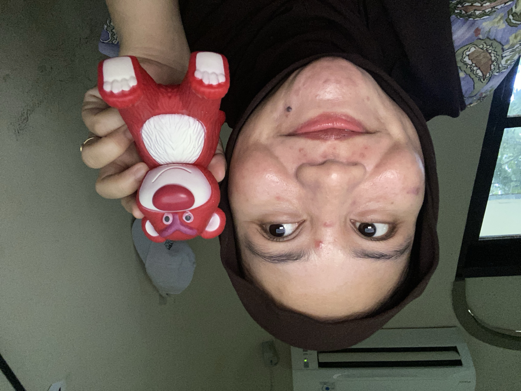 The media file is a selfie taken by a person wearing a brown hijab. The person is holding a red toy with white accents, which appears to be a Lotso Huggin' Bear character from Toy Story, in their left hand. The toy has two large pink eyes with white circles, a pink nose, and a white chest with lines suggesting fur. The person's face is partially visible, and they are looking towards the camera. Visible skin details on their face include some acne. The background appears to be an indoor setting, possibly a room, with a light-colored wall on the left and a window with green foliage visible on the right. An air conditioner unit is visible on the wall below the window. The lighting suggests it is daytime. The overall tone of the image is casual and personal, consistent with a selfie.