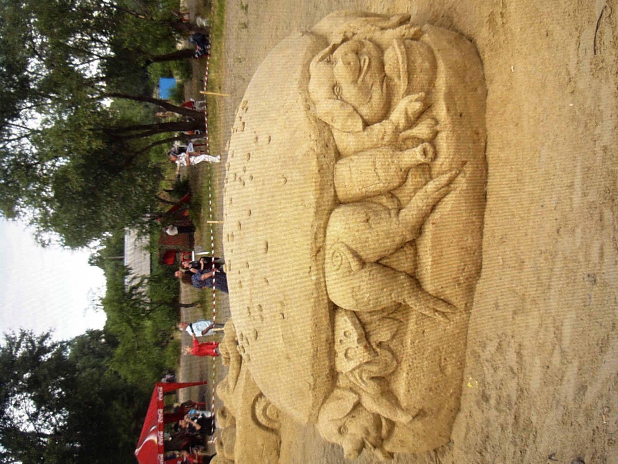 The image depicts a sandy sculpture of a giant hamburger with pigs inside. The sculpture is situated outdoors on a sandy area, with trees and a red tent in the background. Several people are visible in the background, some standing and observing the sculpture, while others are gathered under the tent. The environment appears to be a park or a public space. There are no specific details in the image that allow for pinpointing the exact location within the city of Kyiv, Ukraine, nor are there any actions or interactions occurring between the figures that are clearly discernible beyond observation of the sand sculpture.