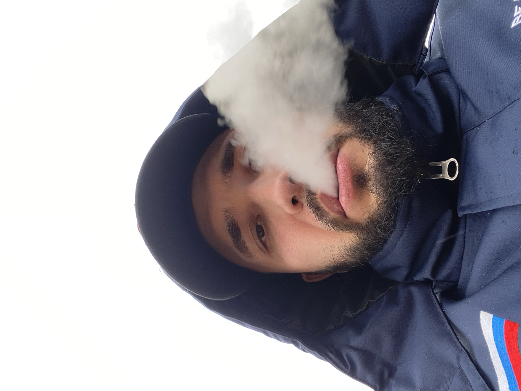 A close-up, low-angle shot depicts a man with a beard wearing a dark blue hooded jacket. The man is exhaling a cloud of white vapor, obscuring part of his face. The background is a bright, white sky. A silver zipper pull is visible on the jacket. The jacket has a stripe of red, white and blue colors. The image does not depict the location of Luzhitsy, Russia.