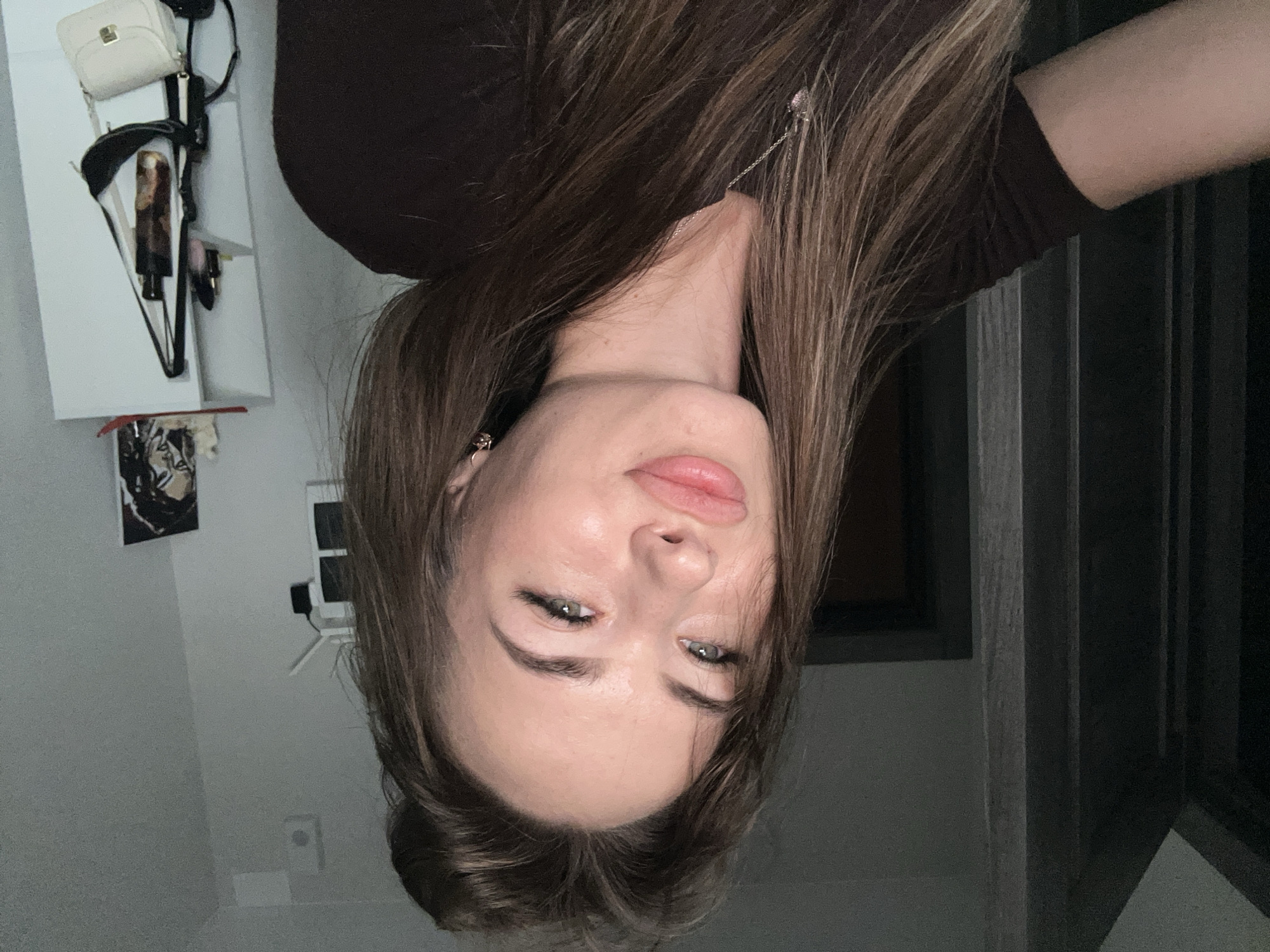 A young woman with long, brown, wavy hair, styled up, poses for a selfie in a dimly lit room. She is wearing a dark brown t-shirt and a silver necklace with a heart pendant. She has full lips, light-colored eyes, and is wearing makeup, including mascara and eyeliner. She is looking towards the camera with a slight pout. In the background, a white shelving unit is visible, with a picture frame, a black bag, and a small white handbag hanging on it. To the left of the frame, a dark doorway and a textured wall are visible. The overall scene suggests a casual, indoor setting.