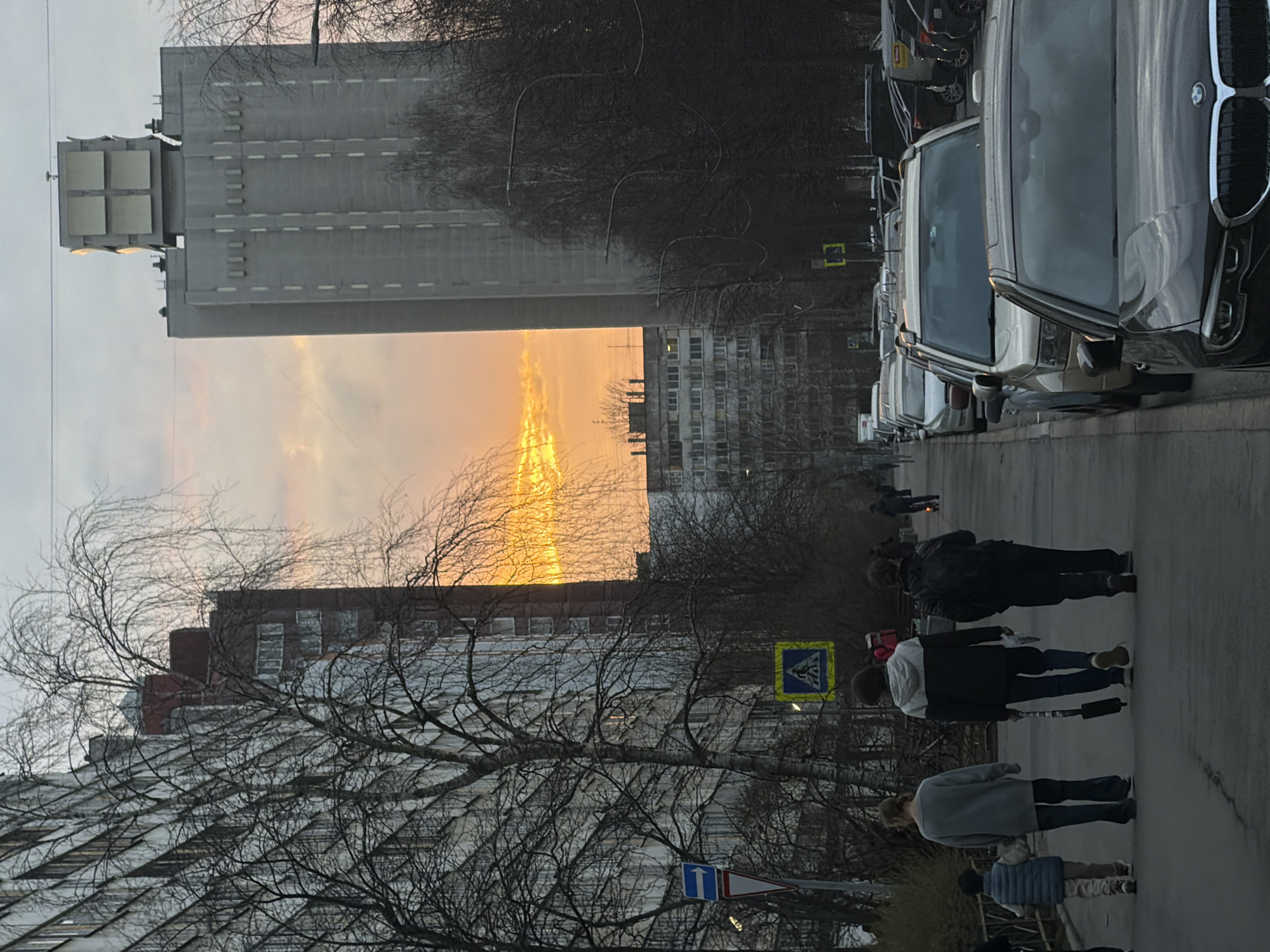 A street scene in Saint Petersburg, Russia. The image is oriented sideways. The street is lined with parked cars on the right side. Pedestrians are walking along the sidewalk. Buildings of varying heights line the street, including one that appears to be a tall, rectangular structure with equipment on top. The sky shows a sunset glow between the buildings. Bare trees are scattered throughout the scene. Traffic signs are visible along the street.