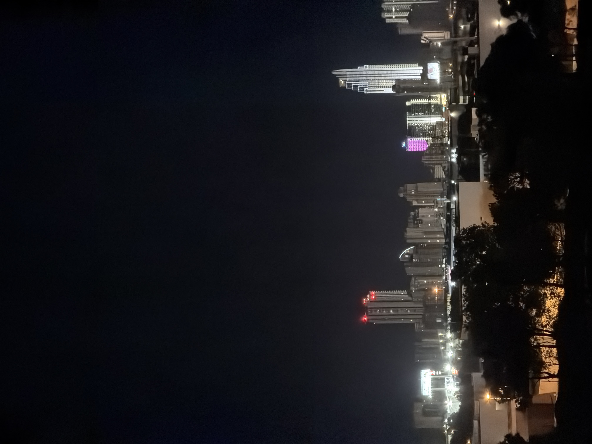 The image shows a nighttime view of the skyline of Finestrat, Spain, with several illuminated skyscrapers. In the foreground, silhouetted trees and foliage obscure the lower portion of the scene. The sky is dark, with the city lights providing the primary illumination. Prominent buildings include a tall, slender skyscraper on the right with a stepped, illuminated facade. To its left, another skyscraper is lit with a pink glow. Further to the left, a cluster of buildings features red lights at their upper levels. In the mid-ground, various other buildings of different heights and designs are visible, some with illuminated windows and signs, including a billboard with the word "ACTION" visible. The overall impression is of a modern, vibrant city at night.
