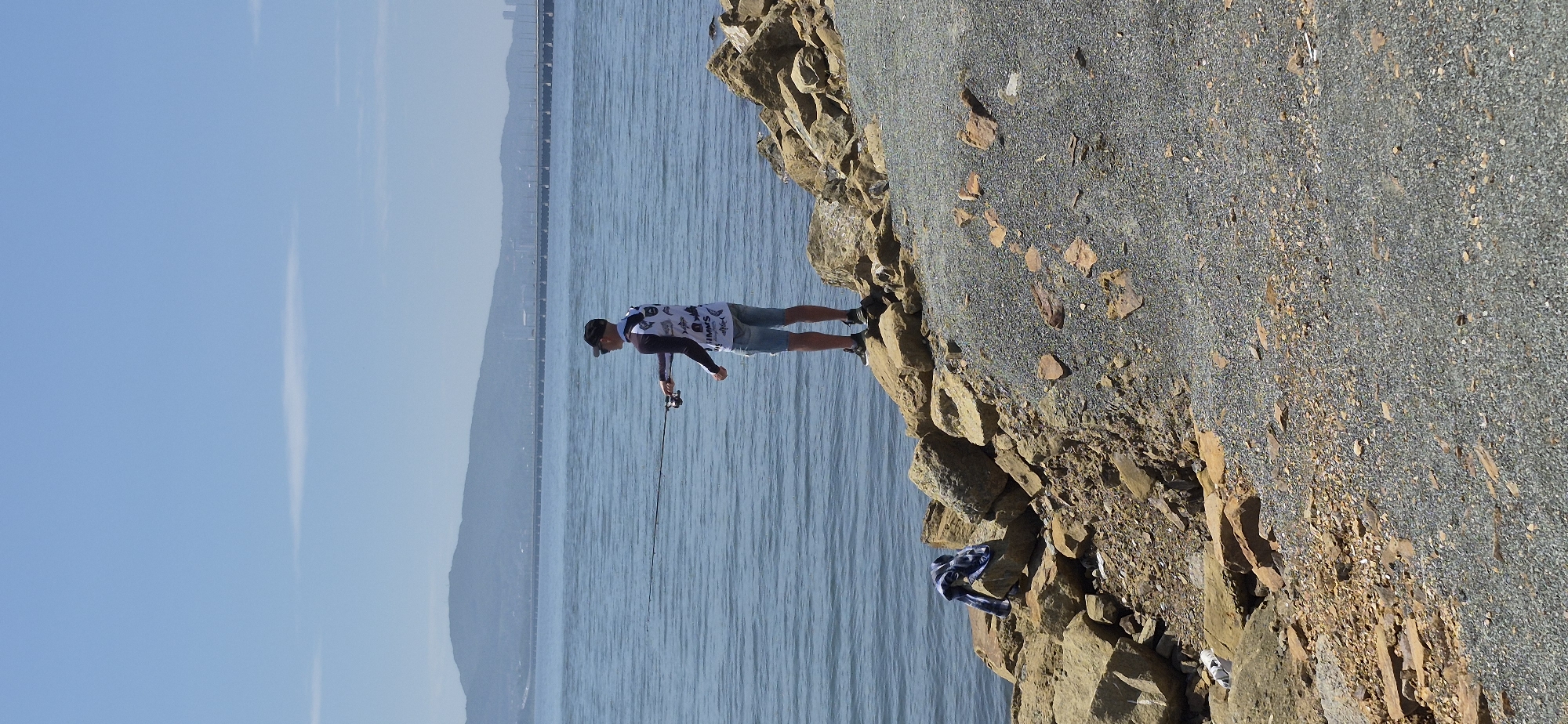 A young man is fishing from a rocky shoreline under a clear blue sky. He is wearing a white fishing shirt with blue graphics, denim shorts, a black baseball cap, and black sneakers. The man is holding a fishing rod and casting into the calm blue water. To his left, a blue and white striped shirt is draped over a large rock. The shoreline is composed of large rocks and gravel. In the distance, a long bridge spans across the water, connecting two landmasses, with a hazy mountain range behind it. The scene appears to be a coastal area, potentially within the city of Vladivostok, Russia, given the presence of a large bridge and mountainous terrain.