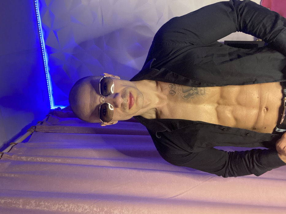An adult male stands posing indoors, directly facing the camera. He is bald and wears octagonal-framed sunglasses with dark lenses. His lips appear slightly colored. He is clad in an unbuttoned black long-sleeved collared shirt, revealing a muscular torso with defined abdominal muscles. A large tattoo depicting a crown is visible on his upper left chest, with a smaller tattoo of "02" positioned above and slightly to the left of the crown. He wears dark trousers with a belt, partially visible at the waist.
The background consists of two distinct sections. To the left, a light pink curtain with vertical folds hangs from a light-colored rod. To the right, a white wall features a three-dimensional geometric pattern. A blue LED strip light runs along the top edge of the wall, casting a blue and purplish ambient light across the scene, particularly affecting the curtain's color. The scene is located in the city of Pleven, Bulgaria.