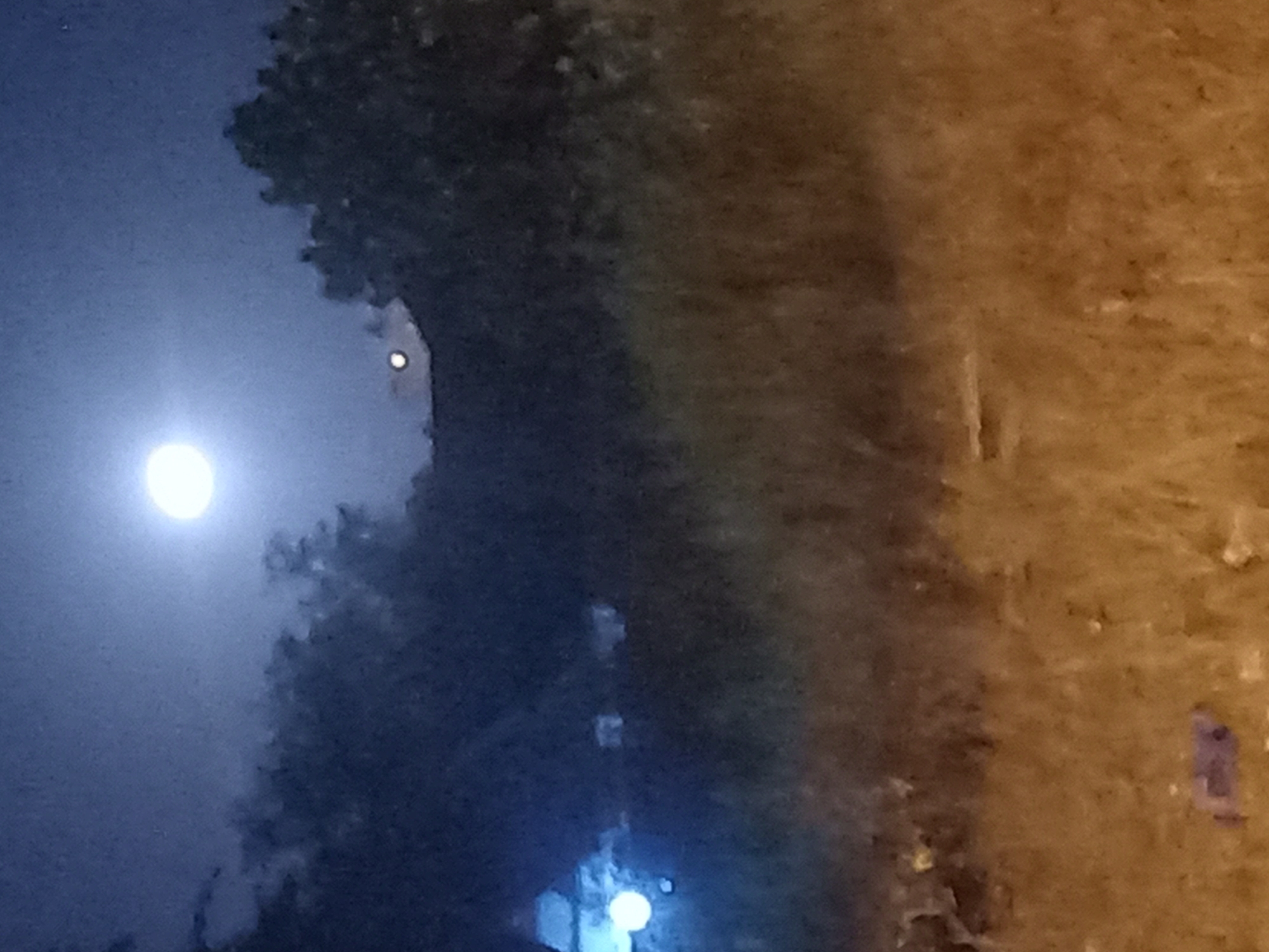 The image appears to be a night scene, possibly in Lhokseumawe, Indonesia. A bright celestial body, likely the moon, illuminates the sky. A dark, undefined mass of foliage or trees is situated between the light source and the right side of the image. In the lower part of the frame, there's a blurred, illuminated area suggesting a street lamp or other artificial light. The right side of the image shows a blurred, textured surface, perhaps a wall or building. The overall impression is one of a poorly lit, nighttime setting.