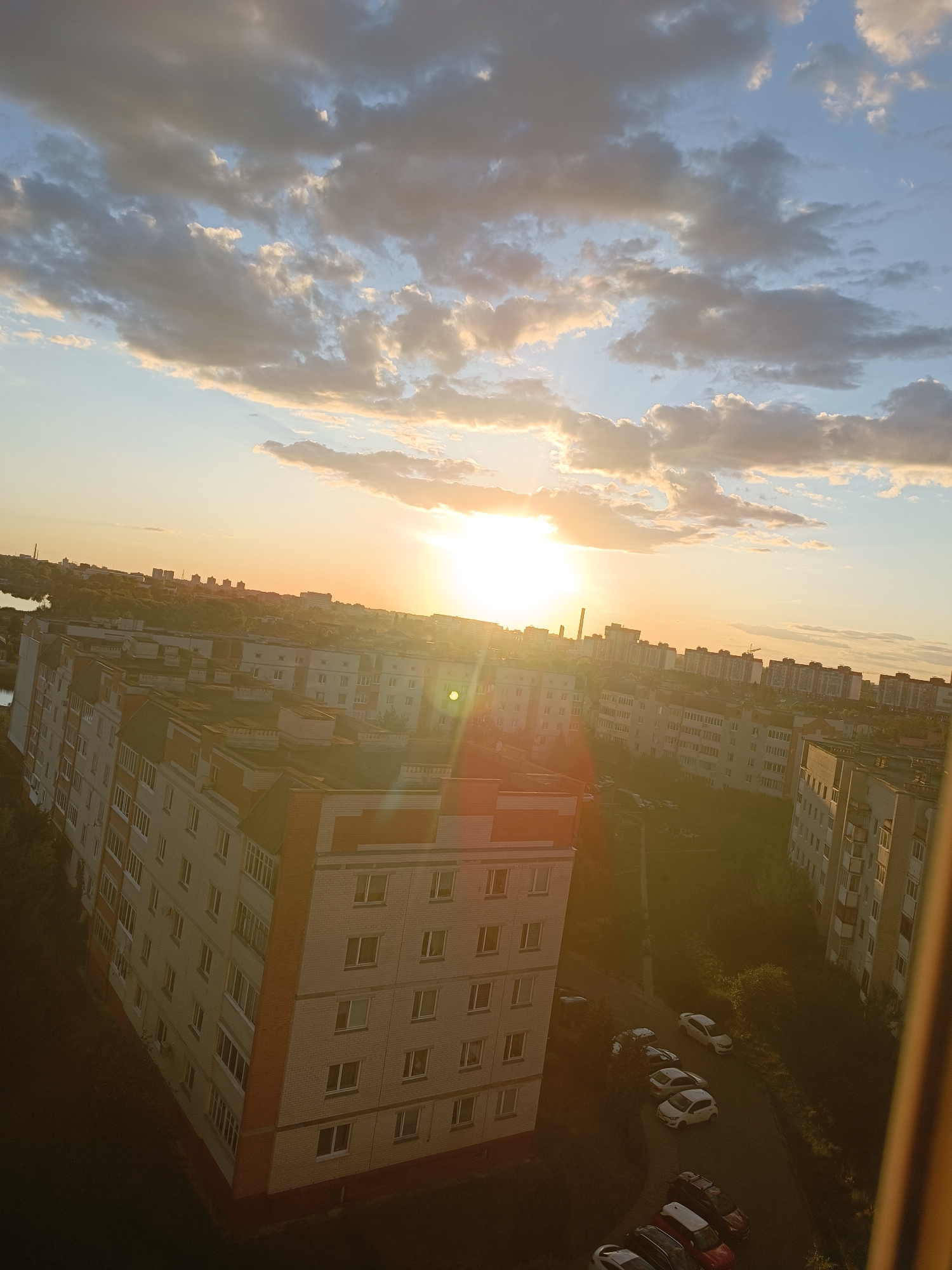 The image captures a sunset over a residential area, likely in Brest, Belarus, as indicated by the architectural style of the buildings. The sun, a bright orb with a noticeable lens flare, is positioned in the upper-middle section of the frame, casting a warm golden light across the scene. Clouds, ranging from light wisps to darker, more substantial formations, are scattered across the sky, reflecting the sunlight.
Below the sky, a panorama of apartment buildings unfolds. The buildings are predominantly light-colored with some sections featuring red brick accents, a common architectural feature in Eastern European cities. They appear to be several stories high and are densely packed together, forming a typical urban landscape. The perspective is from a high vantage point, looking down on the buildings and surrounding environment.
In the foreground, the roofs of some apartment buildings are visible, showing details like ventilation pipes and varying roof textures. Closer to the ground, a street or parking area is visible, with several cars parked. The cars are of various colors, including white, black, red, and silver, and are parked in an organized manner. The presence of trees and green spaces between and around the buildings suggests a degree of urban planning that incorporates nature.
To the left of the frame, a body of water, possibly a river or canal, can be seen in the distance, reflecting the sky. Beyond the immediate residential area, a more distant cityscape with taller buildings and industrial structures is faintly visible against the horizon. The overall atmosphere is serene, with the golden hour light creating a peaceful, end-of-day mood. No people are visible in the image.