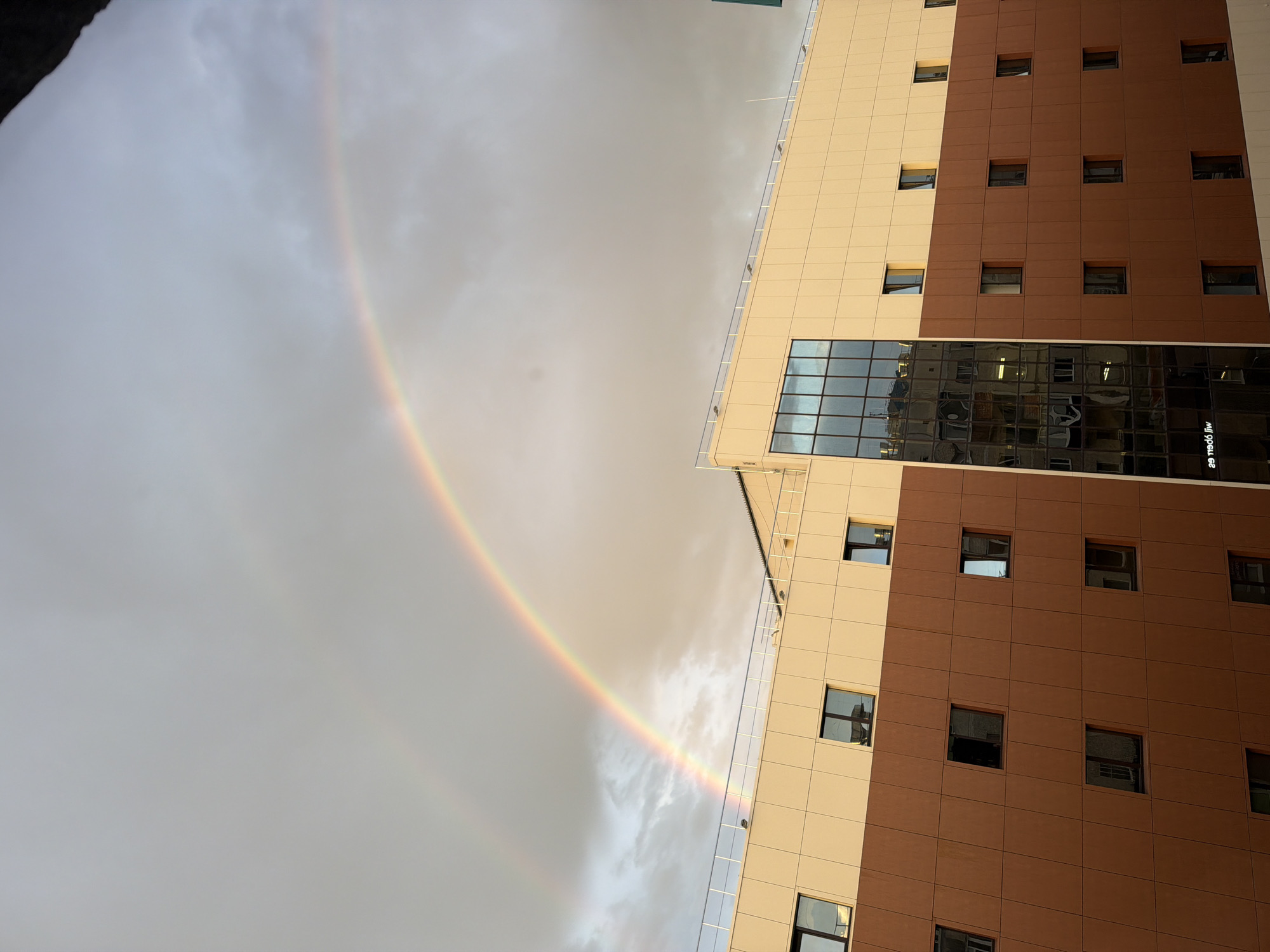The image captures a double rainbow arcing across a cloudy sky above a modern building. The building features a facade with two distinct sections: a lower portion clad in a reddish-brown material and an upper portion in a light beige. Numerous rectangular windows are visible across both sections of the building. A large, dark-paned glass section is present on the upper beige portion, reflecting the sky and the surrounding environment. A white railing runs along the edge of the roof. The scene appears to be an urban setting, likely in Surgut, Russia, given the context. There are no people visible in the image. The overall atmosphere is one of dramatic natural beauty juxtaposed with contemporary architecture.