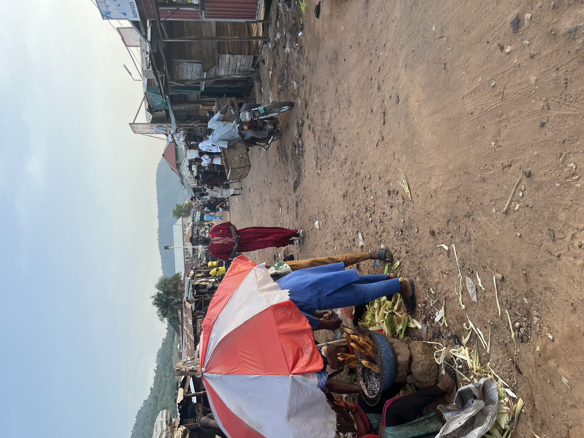 The image depicts a bustling outdoor market scene in an urban environment. The ground is covered in dirt and debris, with tire tracks visible. Along the sides of a dusty path, numerous stalls are set up, constructed from corrugated metal and wood, displaying various goods. In the foreground, a vendor stands under a red and white umbrella, attending to a grill where corn is being roasted. Several ears of corn and their husks are scattered around the stall. People are walking along the path, some appearing to be shoppers and others vendors. One man, dressed in a traditional long red garment, walks past the corn stall. Further down the path, a man is riding a motorcycle with a large wooden crate secured to the back. The background shows buildings and hills under a clear sky. The overall impression is one of a lively and active marketplace.