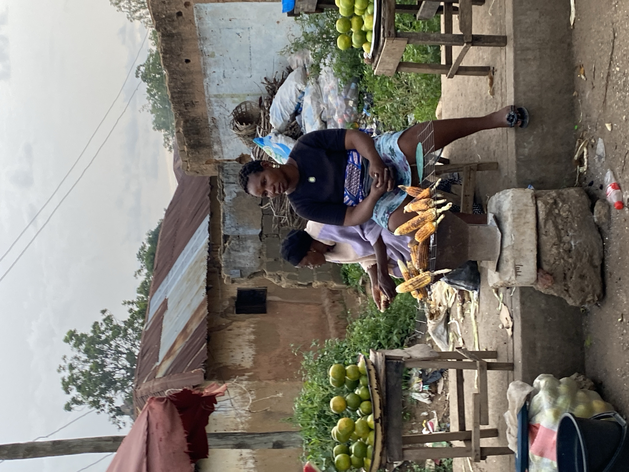 The image depicts an outdoor market scene in Bukuru, Nigeria. Two women are present. The woman in the foreground, wearing a black t-shirt and a patterned blue and white skirt, is seated and appears to be tending to grilled corn. She has short hair and is wearing earrings. Her hands are visible, holding a piece of corn. To her left, another woman, wearing a light purple garment and a dark headscarf, is also seated. She is holding a piece of corn and looking towards her right.
In front of the seated women, there are several ears of grilled corn arranged on a metal rack over a cooking vessel. To the left, a display of green oranges is stacked on a metal tray, supported by a wooden table. Another wooden table with oranges is visible on the right side of the frame. The background shows a collection of structures with corrugated metal roofs and walls made of various materials, including mud and possibly concrete blocks. Piles of bags and debris are present behind the women. The ground is rough and appears to be made of dirt and concrete. The sky is overcast.