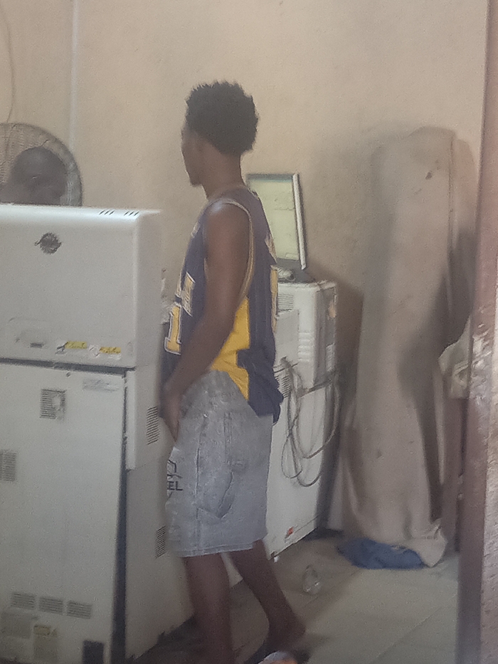 The image depicts an indoor scene in Akwanga, Nigeria. A male individual with dark, curly hair stands facing right, away from the camera. He is wearing a dark blue and yellow sleeveless jersey over light-colored denim shorts. His hands are positioned near his waist. To the left, a tall, white, two-part appliance, resembling stacked commercial refrigerators or freezers, is visible. An oscillating fan is partially obscured behind the upper portion of this appliance. To the right of the individual, a white cabinet supports an active computer monitor displaying a graphical interface, with multiple cables draped over the cabinet's front. Further right, a tall object covered with a light-colored sheet stands against the wall. A blue fabric item and a clear plastic bottle are present on the floor in the foreground. The walls are light-colored and show signs of wear.