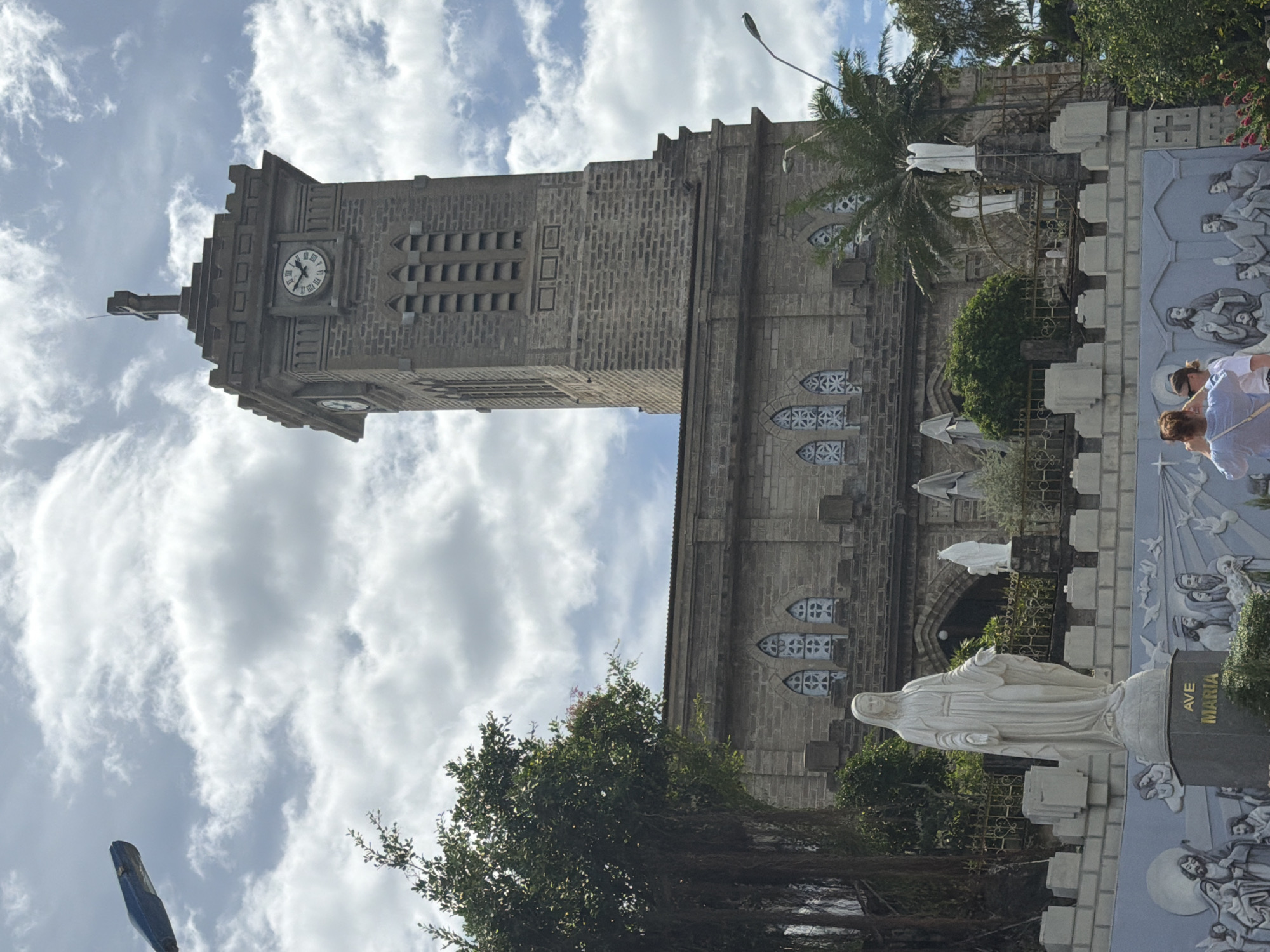 The image shows the Nha Trang Cathedral in Nha Trang, Vietnam. The cathedral is a Gothic Revival-style Catholic church built of stone. The main facade features a tall bell tower with a clock face and a cross at the top. Below the tower, the main body of the church has arched windows with decorative stonework. In front of the church, there is a statue of the Virgin Mary, with other statues and a decorative mural depicting biblical scenes. Two people are visible in the foreground, taking pictures. The sky is partly cloudy, and some trees and palm trees are visible in the surroundings. The image is taken from a slightly low angle, looking up at the cathedral.