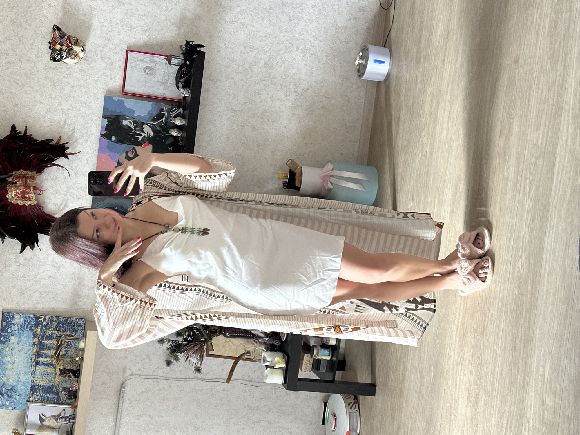 A young woman stands taking a selfie in a brightly lit indoor setting, likely a bedroom or living area. She has long, dark reddish-brown hair and is looking directly at the camera with a subtle smile, her hand near her chin. She is dressed in a white slip dress, covered by a long-sleeved robe with beige and brown geometric patterns, and wears pink fluffy slippers. Her fingernails are painted pink, and her toenails, visible through her slippers, are painted a light blue-green. She also wears a multi-stone necklace and a ring on her left hand.
The background reveals an eclectic room decor. The walls appear to have a light, textured wallpaper, and the floor is a light wood-look laminate. To the woman's left (viewer's right, if image were rotated), a Venetian-style harlequin mask hangs on the wall near a large, red-feathered headdress. Below these are a painting featuring a figure with a mask (resembling Batman), a framed drawing, and a black shelf displaying various small decorative items, including dark feathers and figurines. Further down, another painting with blue and yellow impressionistic tones is visible, along with a shelf holding a deer head sculpture and green foliage.
To the woman's right (viewer's left), a white-and-beige striped basket sits next to a small black table or shelf adorned with candles and other trinkets. On the floor nearby is a light blue cylindrical gift box tied with a pink ribbon. In the upper right corner of the scene, a white and blue humidifier or air purifier is mounted on the wall, with a black cable running from it. A white robotic vacuum cleaner is visible on the floor in the lower right. The overall impression is a personalized and comfortable space, well-decorated with art and unique items. No visible text is clearly legible in the image.