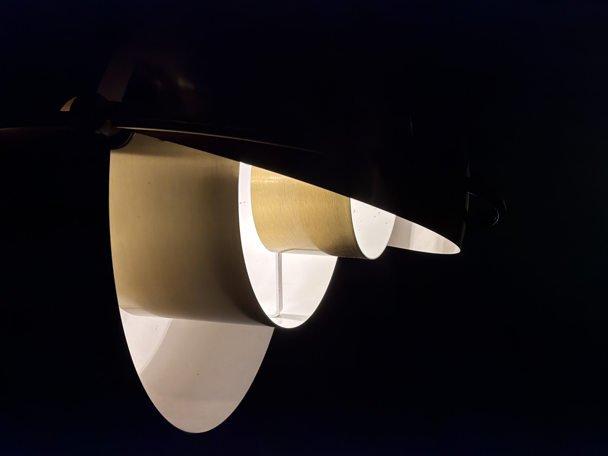 The image displays a close-up of an illuminated, modern light fixture against a completely dark, featureless background. The primary subject is the light source itself, which appears to be a pendant lamp or a wall sconce with a distinctive, multi-layered design.
The fixture is composed of several broad, curved shades. The outer surfaces of these shades are a light, almost white or off-white color, while the inner, illuminated surfaces exhibit a warm, golden, or brass-like hue. A subtle brushed metallic texture is visible on these inner surfaces where the light catches them. The design features overlapping, curvilinear elements that create a sculptural form, guiding and diffusing the light emitted from within. The light source itself is hidden, allowing the light to glow softly around the edges and through the gaps of the shades.
The scene is an indoor setting, likely a room or a display space, but the dark background effectively isolates the lamp, making it the sole focal point. The darkness suggests either nighttime, a very dim room, or an intentional artistic choice to highlight the lamp's design and illumination. There is no visible activity, people, or other objects in the frame. The image emphasizes the lamp's elegant form and the soft, ambient light it produces, evoking a sense of modern aesthetic and warmth. No text is visible.