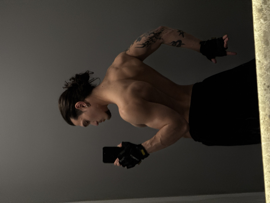 A male individual is depicted from the back and left profile in a mirror selfie. He possesses a muscular physique, with a bare torso exposing his back, shoulders, and arms. His dark hair is styled in a man bun. He has a trimmed beard and mustache, and a silver hoop earring is visible in his left ear. Multiple dark-colored tattoos, including what appears to be a snake and stylized elements, cover his left arm (viewer's right). He wears black fingerless gloves on both hands and black shorts or sweatpants. A black smartphone is held in his right hand (viewer's left), angled towards the mirror, indicating he is taking the photograph. His left hand (viewer's right) is held open with fingers extended. The background is a plain, solid grey wall, reflected in the mirror. A narrow strip of a light-colored, possibly stone or tiled, surface is visible at the bottom of the mirror's frame or reflection, likely a countertop. The lighting is artificial and highlights the subject's musculature. No identifiable features within the image indicate a specific location in Sofia, Bulgaria; the setting appears to be a generic interior space.