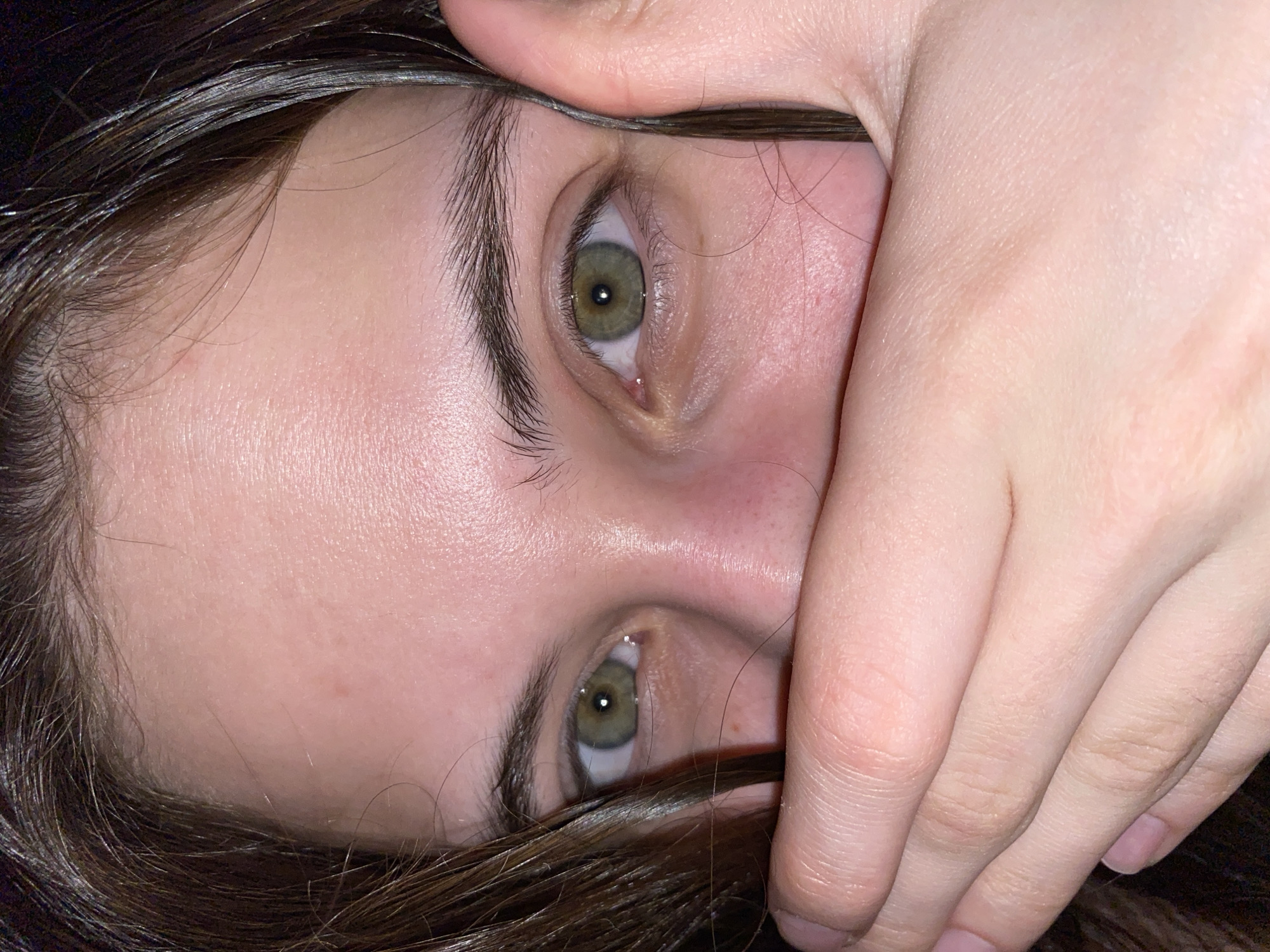 A close-up, upside-down view of a person's face. The person has fair skin, brown hair, and green eyes. A hand is partially covering the person's face, obscuring the mouth and nose. The location is not discernible from the image.