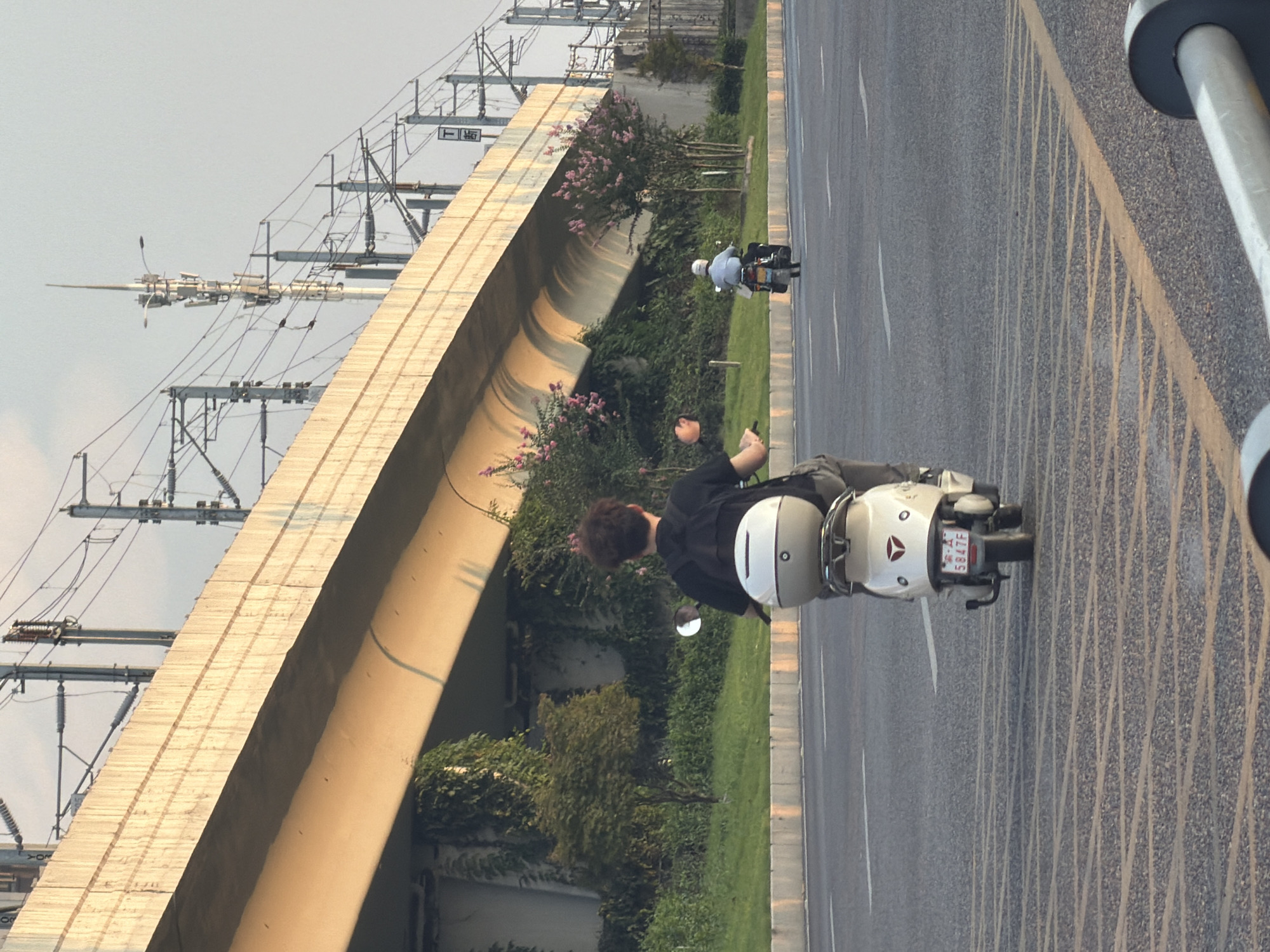 The image depicts a scene in the city of Jiangjin, China, featuring two individuals riding scooters on a road under a large elevated highway. In the foreground, a person wearing a black t-shirt and a backpack is riding a white scooter with a white top box. The scooter has a red license plate with the characters "A 5847F". In the background, another person is riding a motorcycle, further down the road. The environment includes the highway overhead, with its concrete structure and supporting power lines and poles. Green vegetation, including trees and bushes, grows along the side of the road beneath the highway. The sky is visible above, with a hazy or overcast appearance. The road surface is asphalt, marked with white and yellow lines. The lighting suggests it might be late afternoon or early morning, with the sun casting a warm glow on the highway structure.
