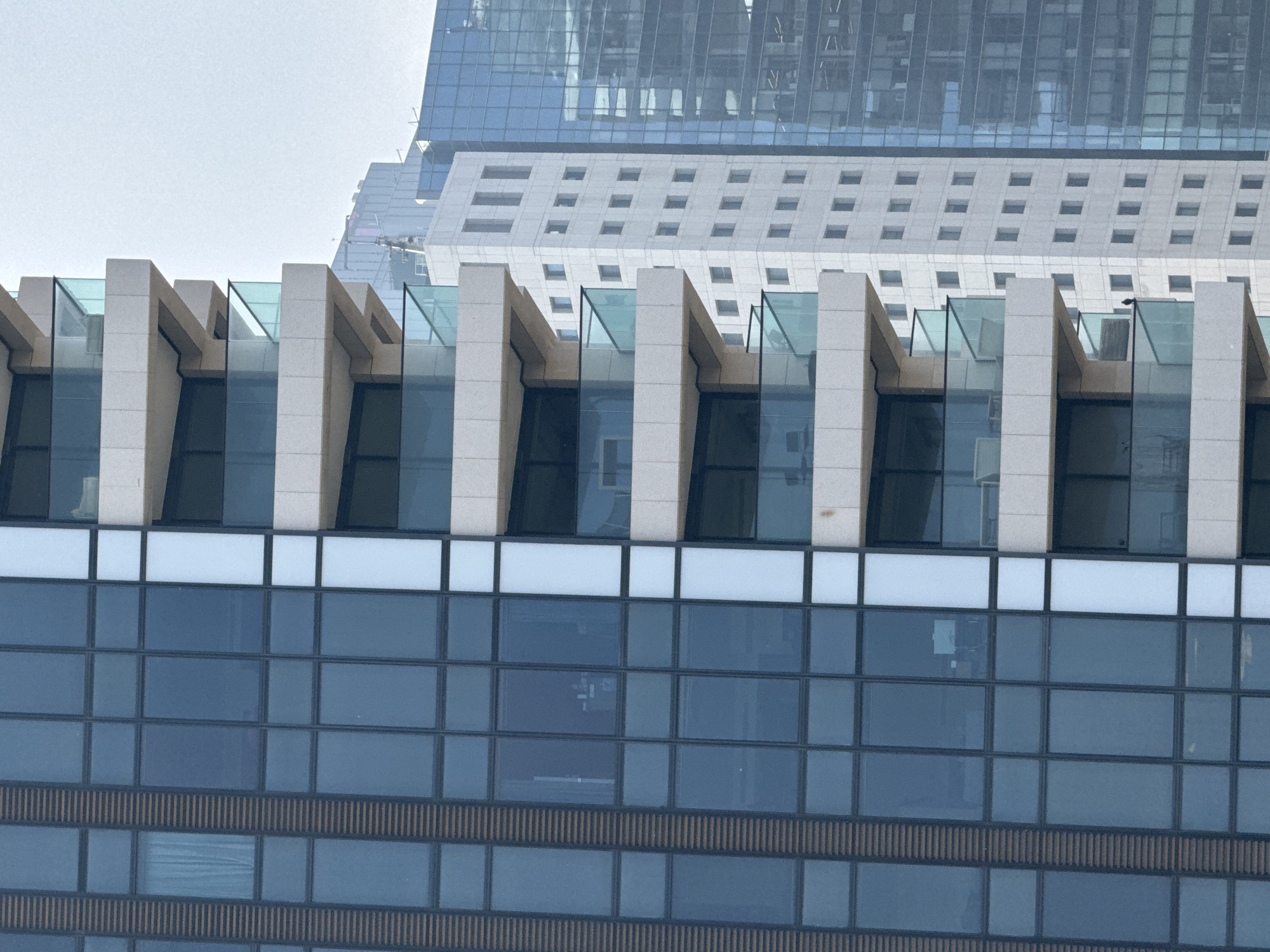 The image captures a close-up view of building facades in Tel Aviv, Israel. In the foreground, the top portion of a dark, blue-windowed building is visible, featuring a white rectangular division above the windows. Above this, a distinct row of evenly spaced, cream-colored structural supports with glass paneling can be seen. In the background, another building is partially visible with rows of regularly spaced square windows and a larger, glassy structure rising above it. The sky in the background is light gray, suggesting overcast conditions.