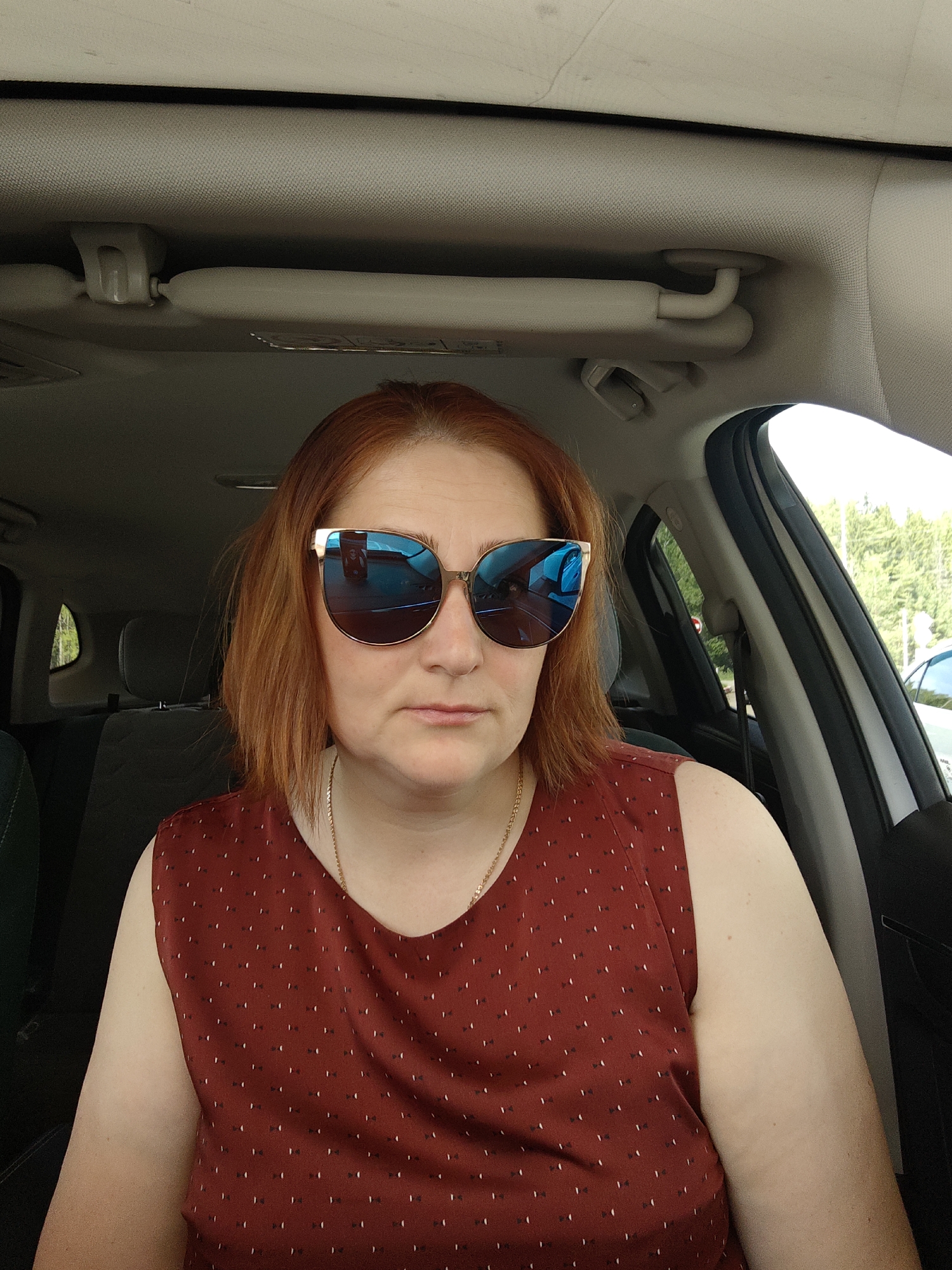 A middle-aged woman with red hair is sitting in the driver's seat of a car. She is wearing large sunglasses that reflect the interior of the car. She has on a maroon top with small patterns. A gold necklace is visible around her neck. The interior of the car is visible, including the dashboard, seats, and sun visor. Through the window, trees and another car can be seen, suggesting the car is parked on a road. There is no specific information in the image to confirm the location in the city of Chaikovsky, Russia.