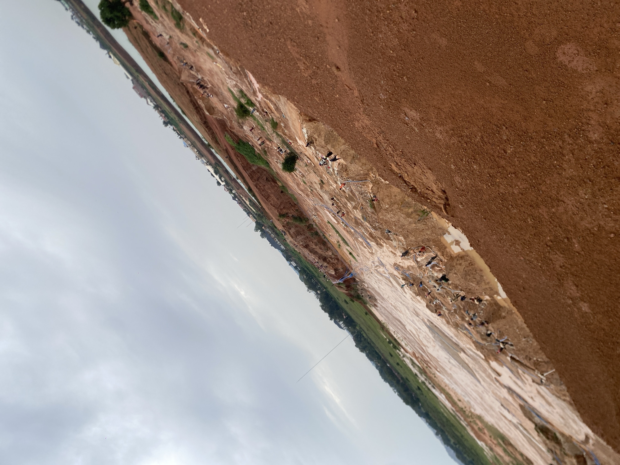 This image depicts a large, exposed reddish-brown earth quarry, likely in Jos, Nigeria, given the context. The quarry floor is uneven and appears to be a mixture of mud and dry earth, with several small pools of water scattered throughout. A significant number of people, appearing to be individuals engaged in mining activities, are spread across the quarry. They are engaged in various actions, including digging, carrying, and moving materials, with some visible interactions among them. Some individuals are clustered together, while others are more isolated. There are visible signs of informal mining operations, including what appear to be makeshift tools and possibly some rudimentary equipment. The surrounding environment shows a contrast between the barren quarry and greener vegetation further away, with a body of water and some buildings visible in the distance, suggesting proximity to a populated area. The sky is overcast with clouds. The image is taken at an angle, giving a sense of depth and the scale of the quarry operation.