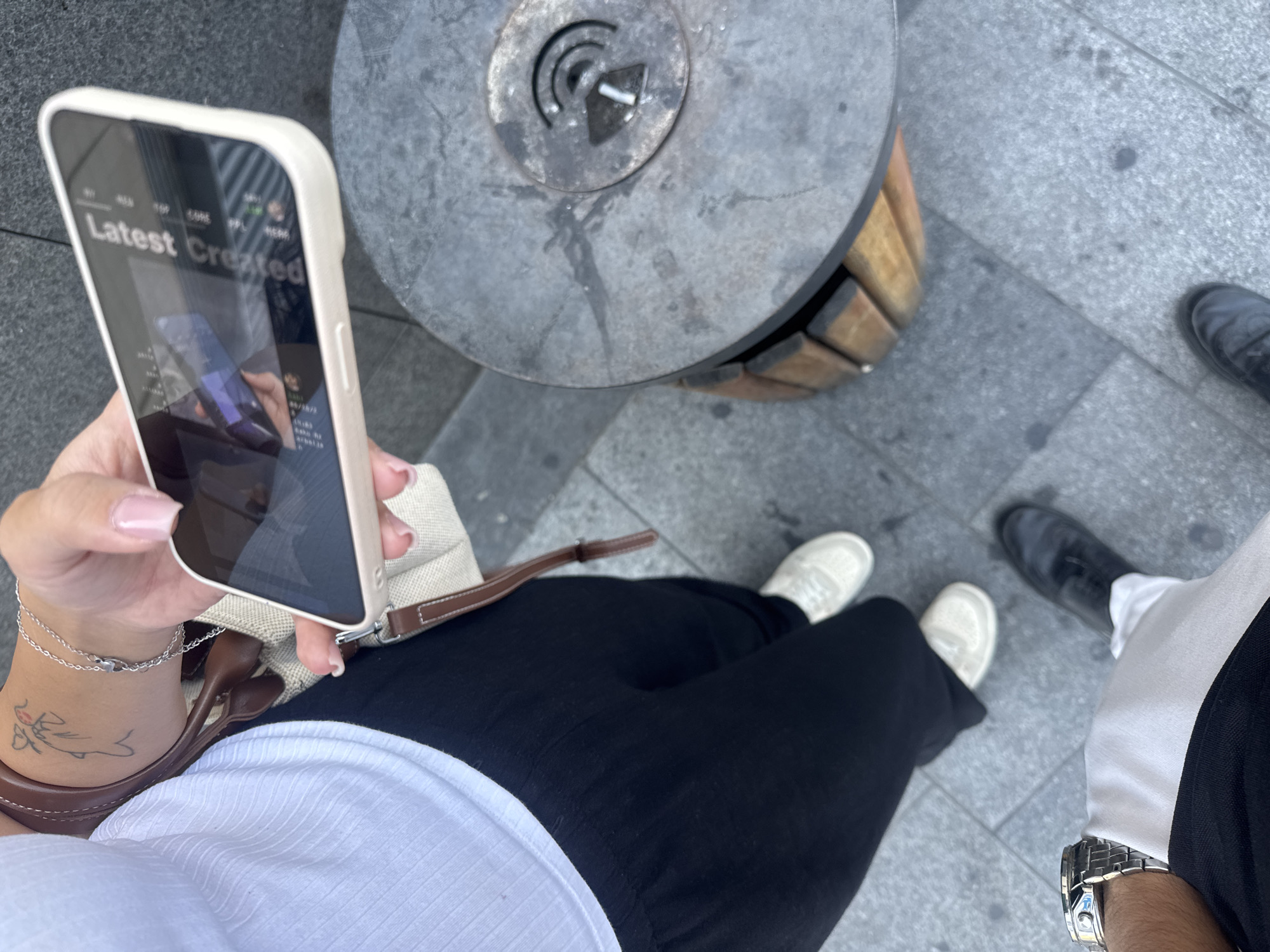 The image shows a close-up view of two people standing on a paved city sidewalk, possibly in Baku, Azerbaijan, as indicated by the presence of a public ashtray with a wireless signal symbol, a common feature in Baku. One person, on the left, is wearing black trousers and white sneakers, with a silver watch on their left wrist. This person is partially visible from the waist down, facing away from the camera. The other person, on the right, is holding a smartphone with a white case in their right hand, displaying a screen with the text "Latest Created". This person is wearing black trousers and white sneakers. The background consists of a gray paved surface and a round metal public ashtray with a wooden base, featuring a symbol resembling a wireless signal. The overall scene suggests an outdoor urban environment.