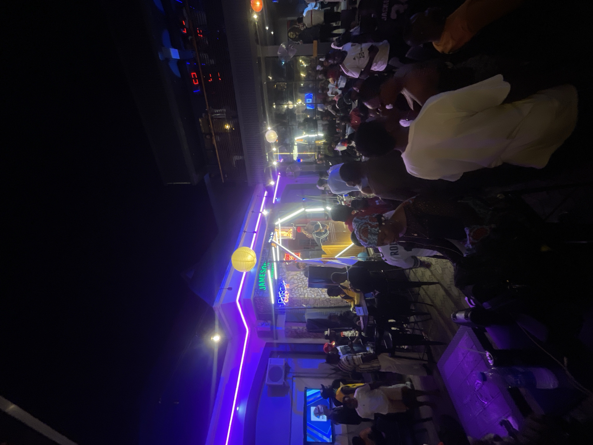 The image shows an outdoor bar in Lokoja, Nigeria, at night. The bar is crowded with people sitting at tables and chairs. The building is lit with blue and purple neon lights and has neon signs for Jameson and Party. There is a television mounted on the wall. The scene appears lively and social.