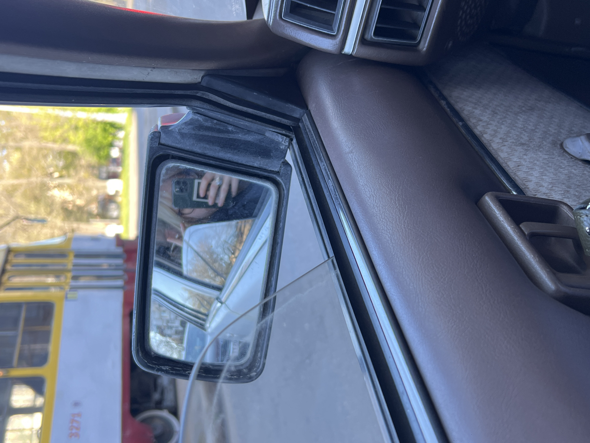 A side mirror reflects a yellow and red trolleybus, specifically trolleybus number 3271, indicating the photograph was taken in Odesa, Ukraine. The reflection also shows a person holding a phone. The mirror is attached to the door of a car. Inside the car is a brown interior and dashboard with vents.