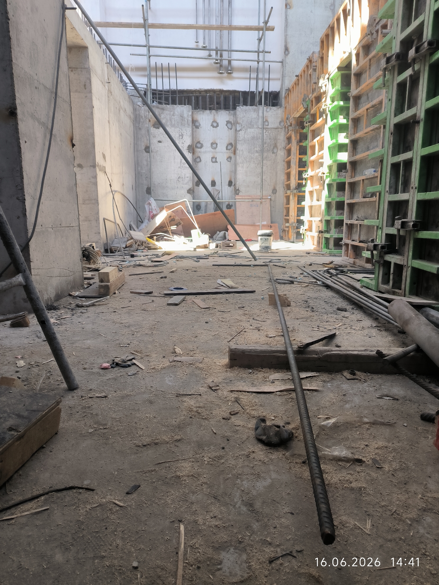 The image depicts an unfinished construction site in an indoor location, possibly a basement or a large room under construction. The scene is messy, with construction debris, including wood scraps, dust, and discarded materials, scattered across the concrete floor. Several vertical rebar rods are visible, some lying on the ground and one prominently in the foreground, suggesting ongoing structural work. On the right side of the image, formwork, likely for concrete pouring, is set up, with sections in orange and green. In the background, concrete walls are visible, along with scaffolding and various construction elements. Sunlight is streaming in from an unseen source, illuminating parts of the scene and casting shadows. The overall impression is one of an active but uncompleted construction project.