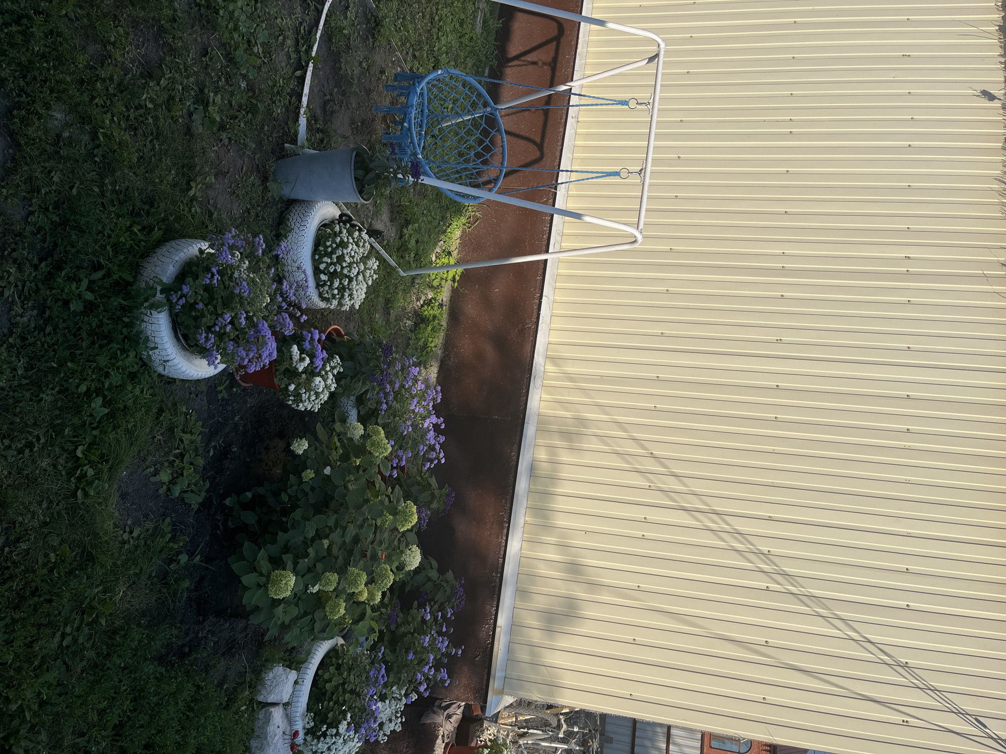 The image depicts an outdoor scene in Saratov, Russia, featuring a swing set with a blue net seat in front of a beige corrugated metal wall. Several potted plants with white and purple flowers are arranged in front of the wall, with some planted in repurposed tires. The ground is covered with grass and dirt. Shadows from an unseen object are cast diagonally across the metal wall. The lighting suggests it is daytime. There are no people or animals visible in the frame.