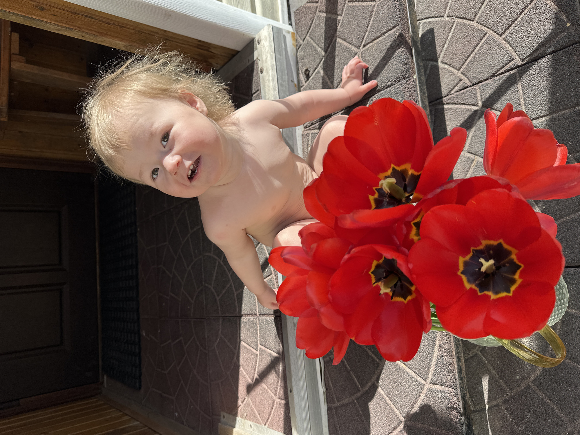 A high-angle shot shows a young, fair-skinned child with light blond hair sitting on a gray tiled surface next to a vase filled with vibrant red tulips. The child is mostly nude, with the flowers partially obscuring the lower part of the body. The child is looking upwards towards the camera with a smile, revealing a few teeth. The background shows a wooden doorway and a white window frame. The scene is lit by bright sunlight, casting shadows on the tiled surface. The location appears to be outdoors, possibly on a patio or porch, but there is no definitive evidence to place it in Nizhny Novgorod, Russia.