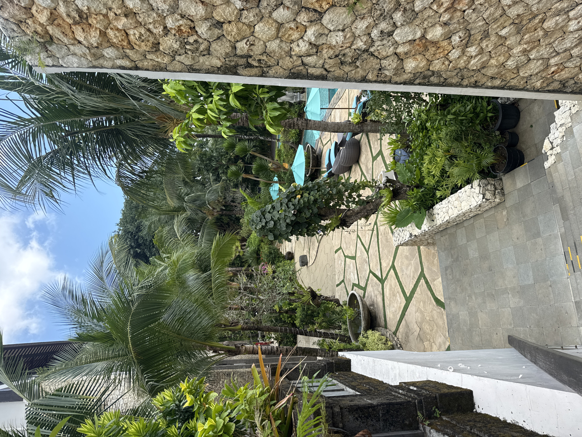 This is a high-angle shot of a resort courtyard in Kuta Selatan, Indonesia. Palm trees and lush green foliage surround a pathway paved with large light-colored stones and accented with green artificial turf. The pathway leads to a swimming pool area with turquoise umbrellas and lounge chairs. A stone wall frames the right side of the image, and a white wall and stairs are visible on the left. The sky is blue with scattered clouds. There are no people visible in the image.