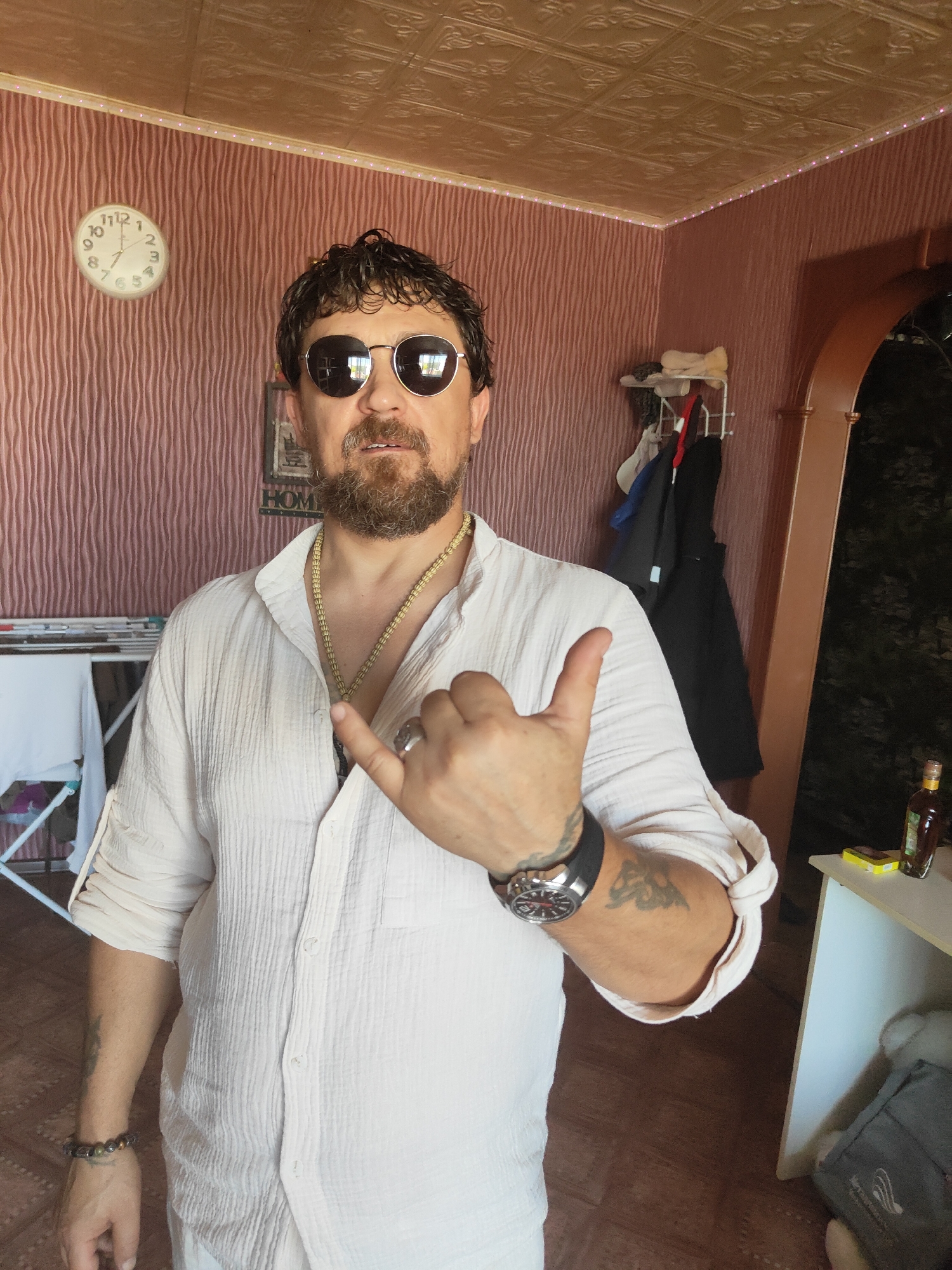 A man is standing in a room, giving a shaka sign. He has dark hair, a beard, and is wearing sunglasses, a white linen shirt, a gold chain, a watch, and bracelets. Behind him, there is a clock on a pink, wavy-patterned wall, a coat rack with clothes, and an arched doorway. A shelf with items is visible to the right. The room appears to be a residential space.