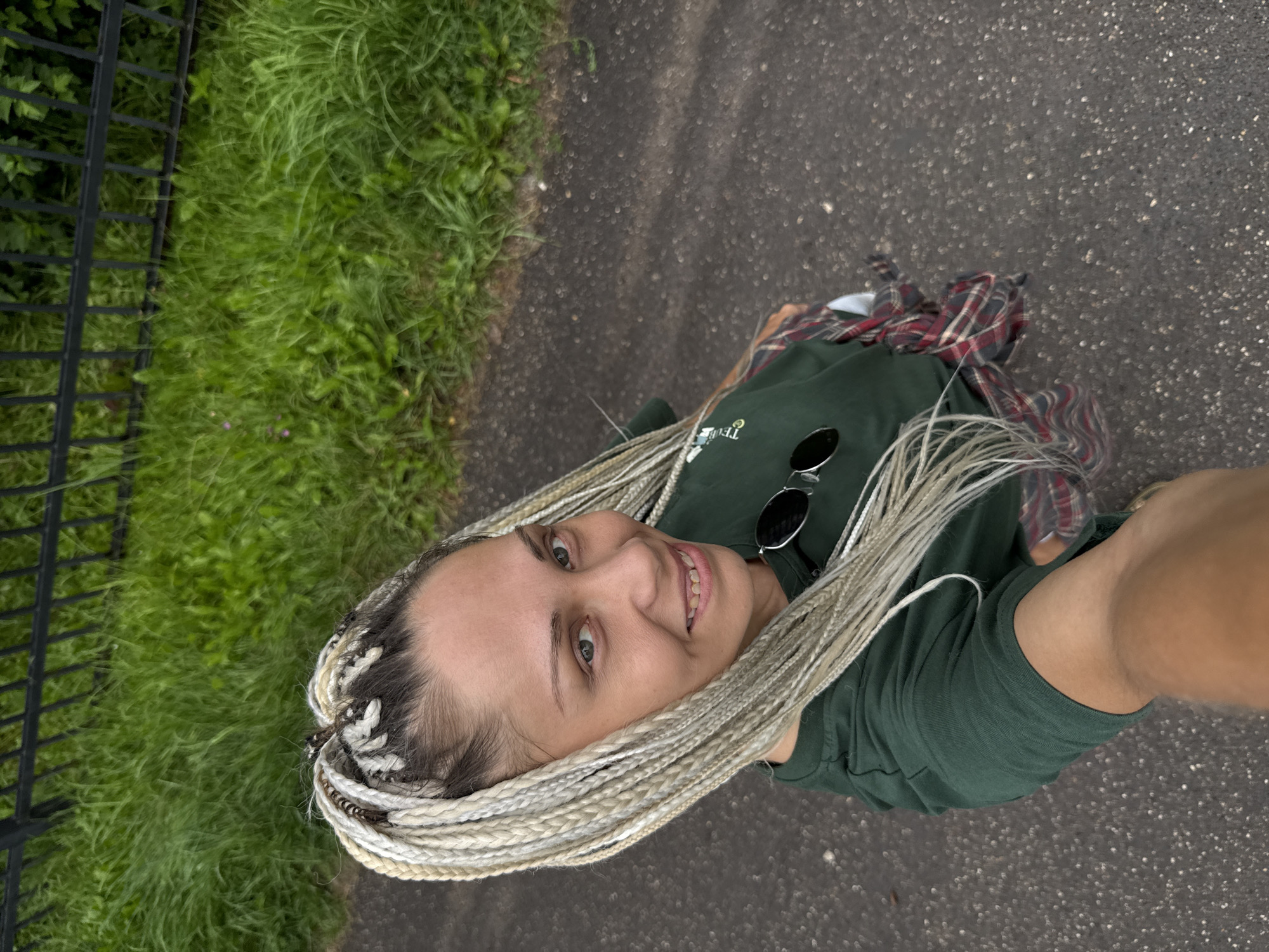 A woman with blonde braids is standing on a paved surface, possibly a sidewalk or path, in Rusanovo, Russia. She is wearing a dark green t-shirt over a red and black plaid shirt tied around her waist. She has on sunglasses perched on her t-shirt. Behind her, a black metal fence runs along a strip of lush green grass. The background suggests an outdoor, possibly urban or park-like setting. The image is taken from a high angle, looking down at the woman.
