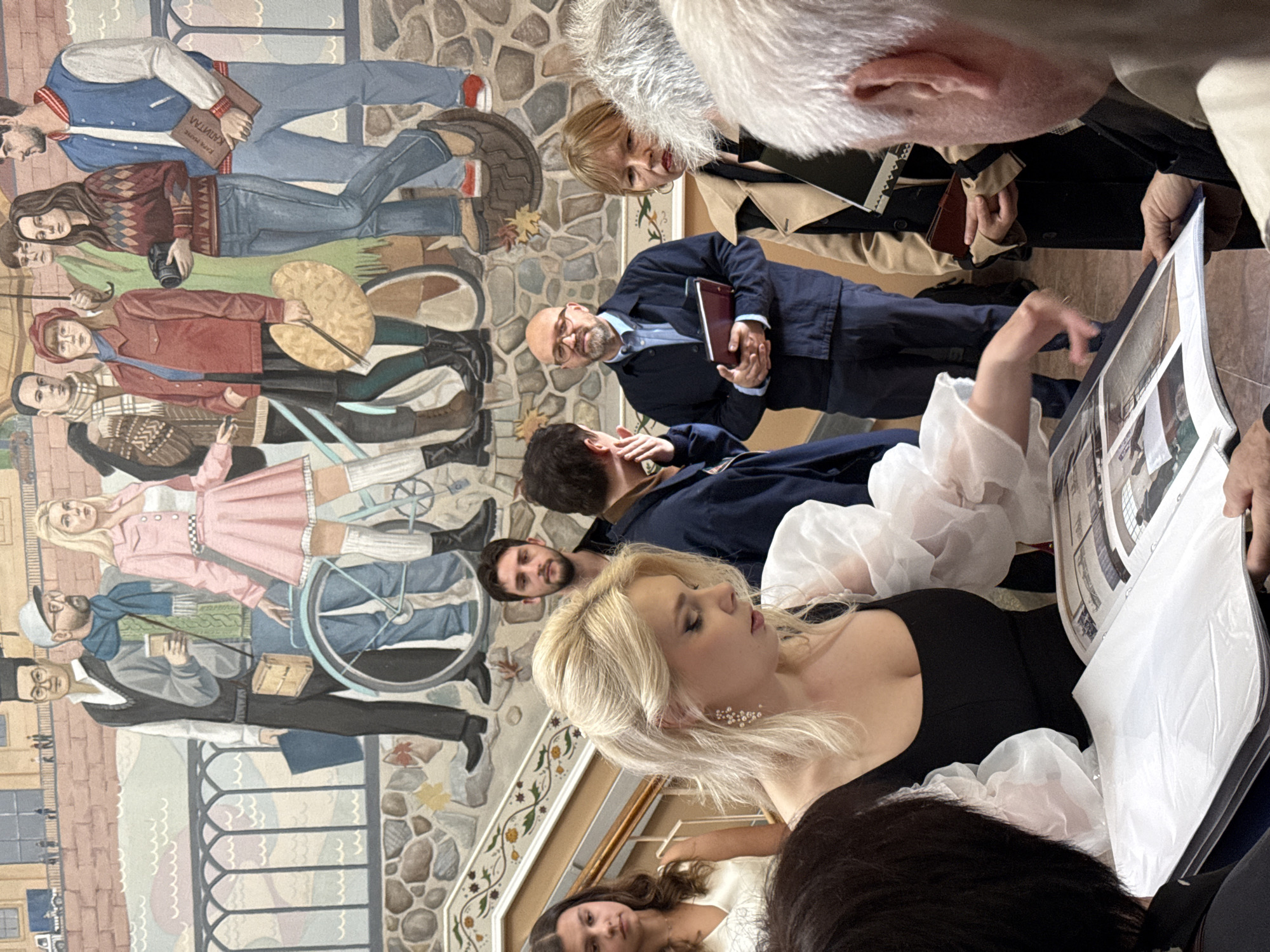 The image depicts a gathering of people in front of a mural on a stone and tiled wall, likely in Saint Petersburg, Russia. The mural features a group of young people in casual, fashionable attire, some holding books, a camera, and art supplies, suggesting a theme of education or artistic pursuit. In the foreground, a woman with blonde hair, wearing a black dress with voluminous white sleeves, is leaning over an open book or portfolio, her hand gesturing as if pointing something out. Several other individuals are present, some looking at the book, others observing the mural or conversing. The environment suggests an indoor or semi-enclosed space, possibly a public building or gallery, with a staircase visible to the left.